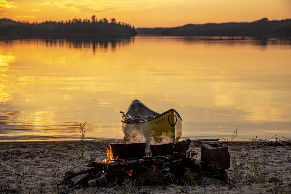 Yellow canoe pulled up on beach in front of campfire at sunset.