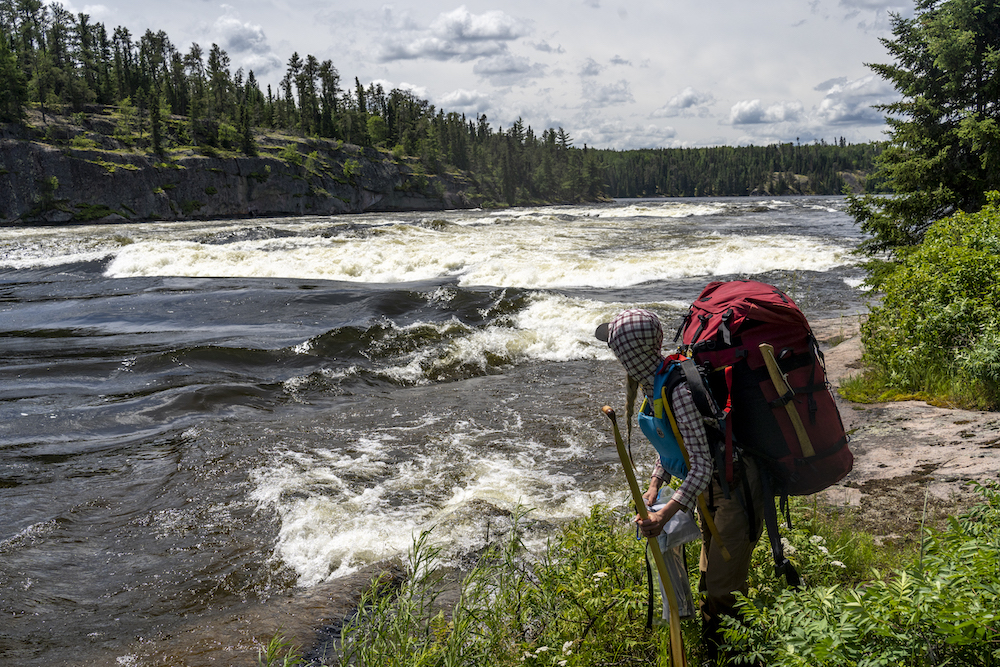 Woman carrying red canoe pack and paddles stands beside rapids.