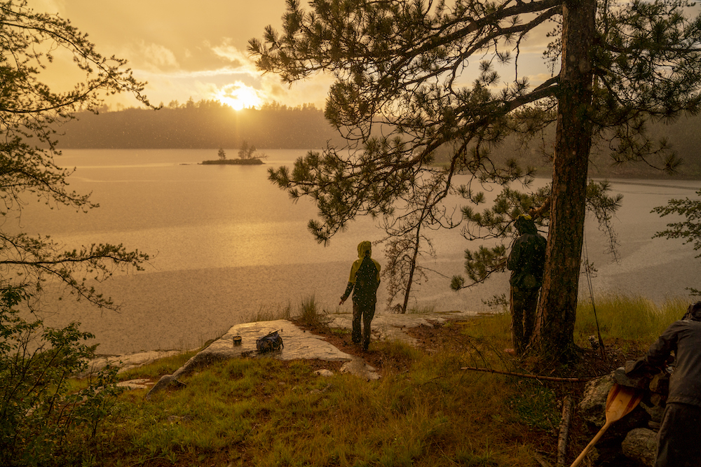 Person stands next to lake as sun peaks out from horizon during rain storm.