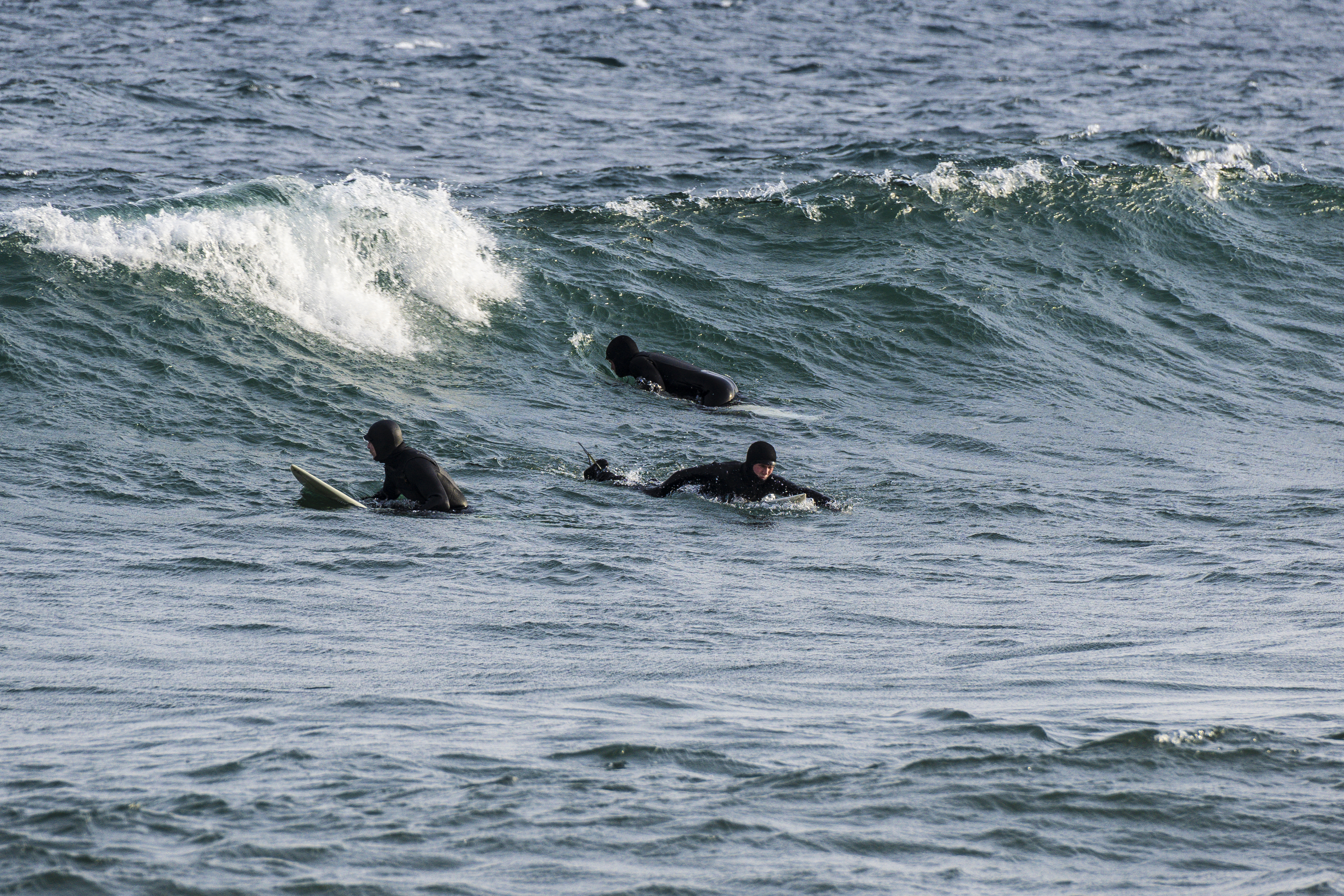 3 surfers in wetsuits paddle to catch a large blue lake wave under a sunny sky