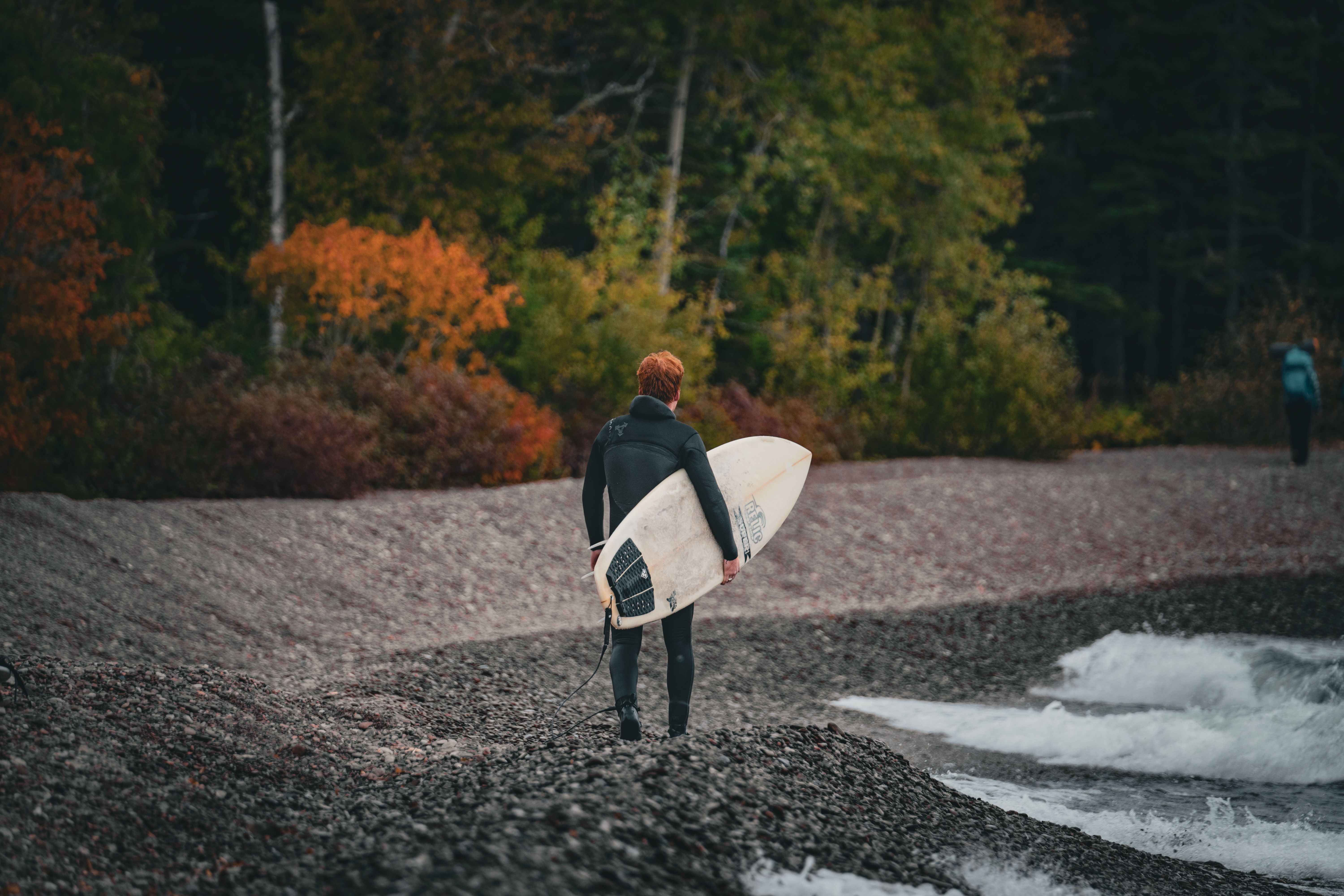 A person in a black wetsuit walking on a rocky lakeshore, holding a surfboard. There are orange and green autumn leaves in the background.