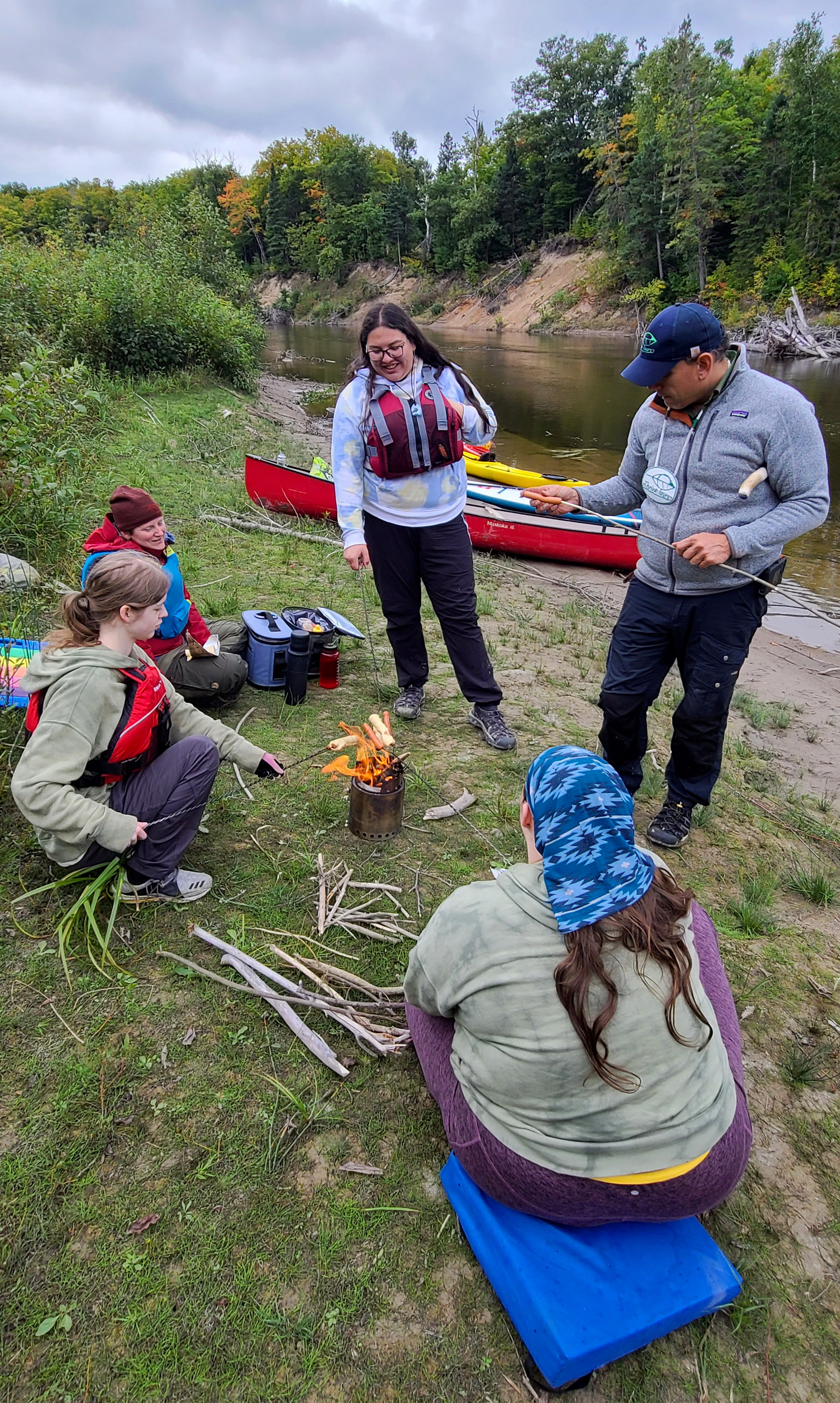 a group of people standing around a small camp fire, talking and cooking on a grassy river bank. Canoes and kayaks sit near the river in the background.