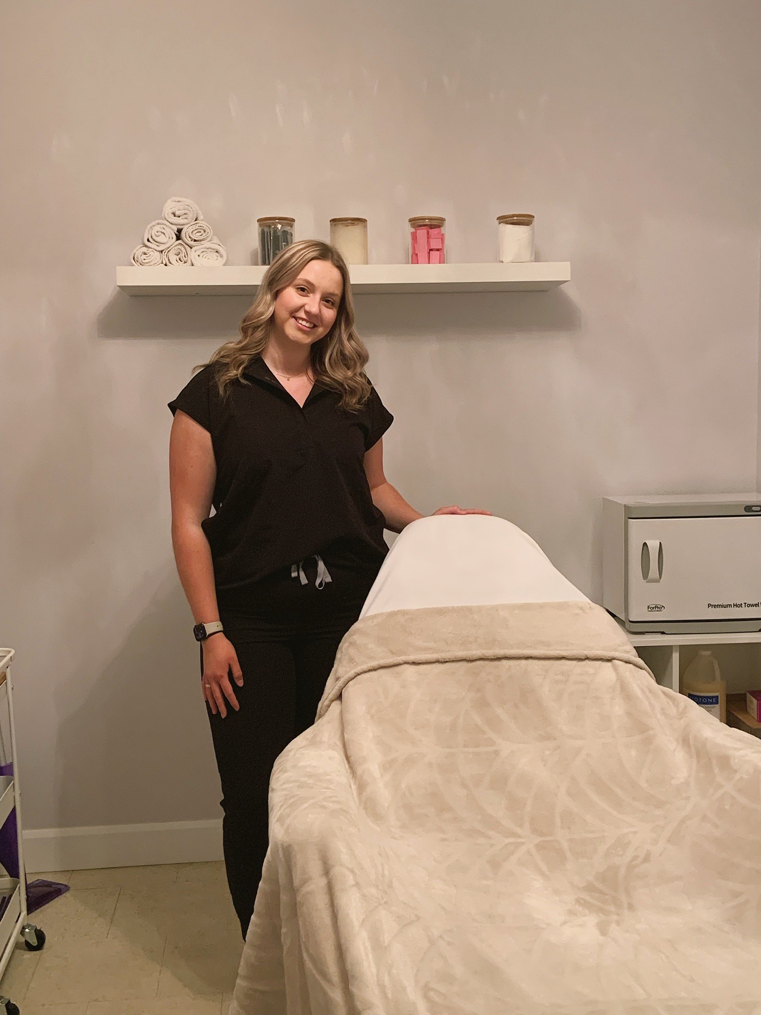 A smiling esthetician standing in front of a comfortable spa bed