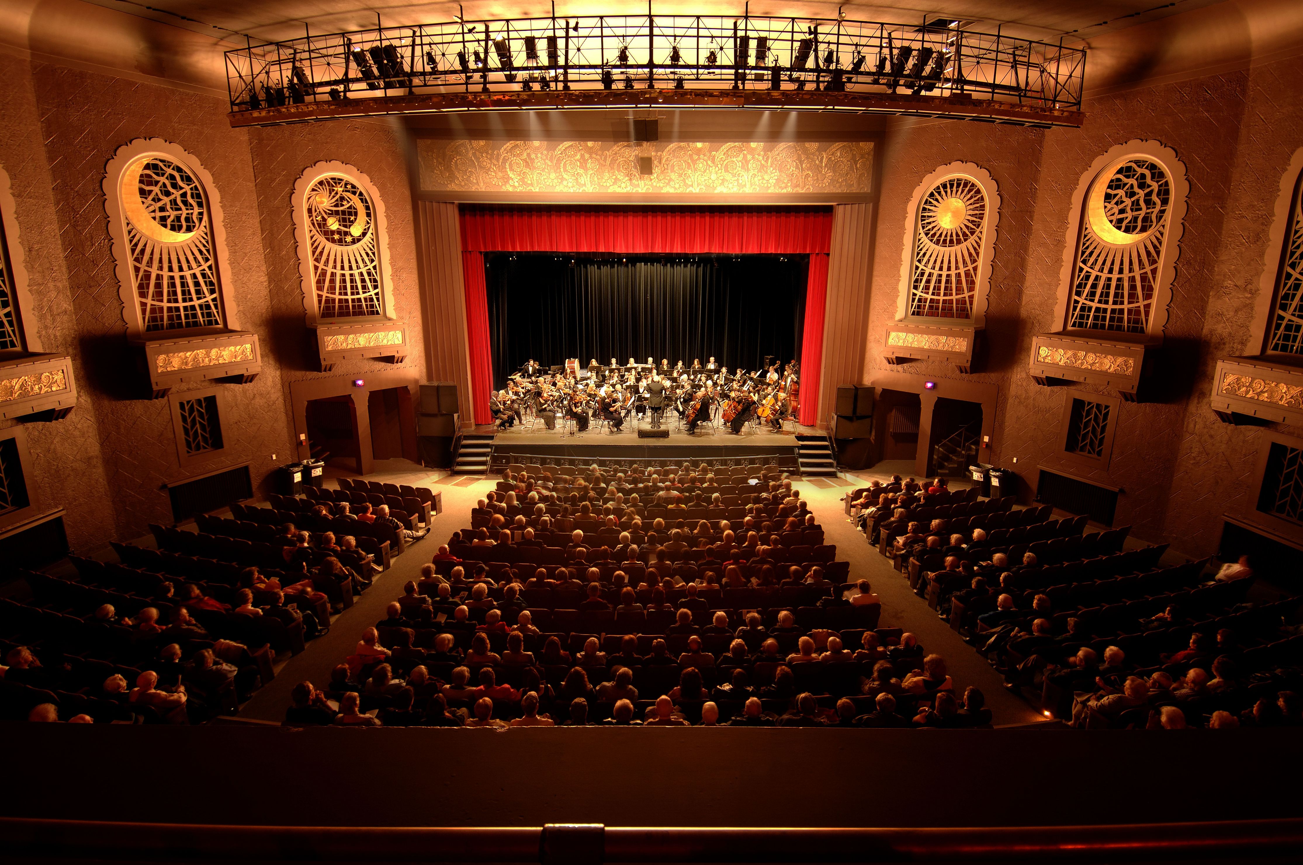 The interior of Capitol Centre in North Bay; a stately old theatre with elegant molded celestial motifs on the walls, and golden spotlights. The rows of seating are filled with audience members and there is an orchestra on stage. 