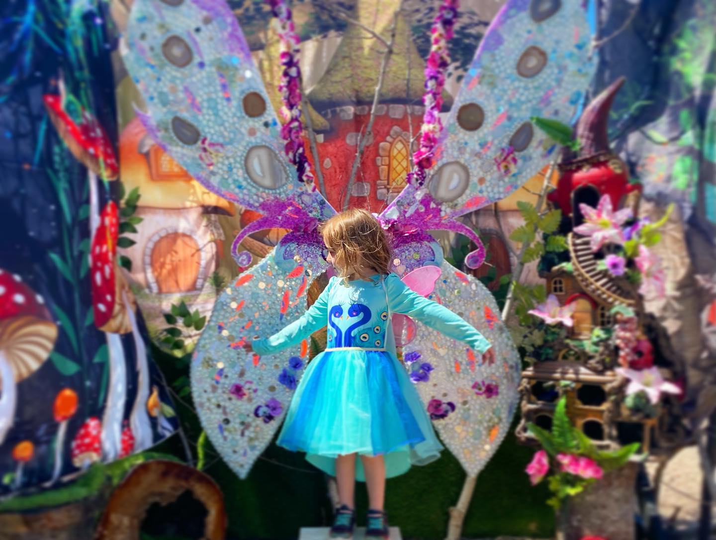 A small girl in a blue dress poses in front of an elaborate and colourful fairy wing display, giving the effect that she has wings.