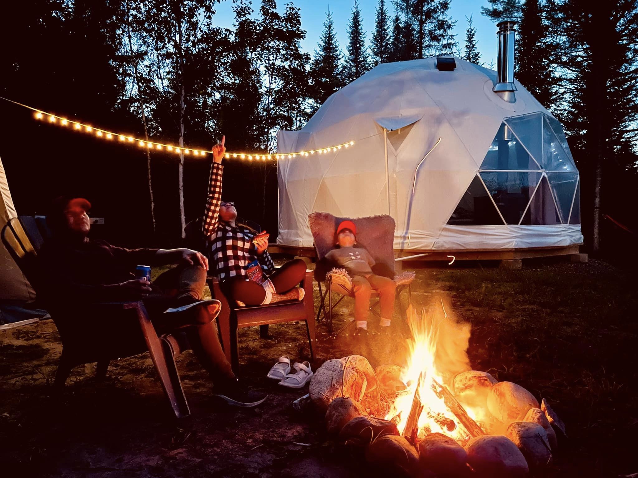 A family sits by a campfire in front of a small geodesic dome. All are looking up and one person is pointing at the stars.