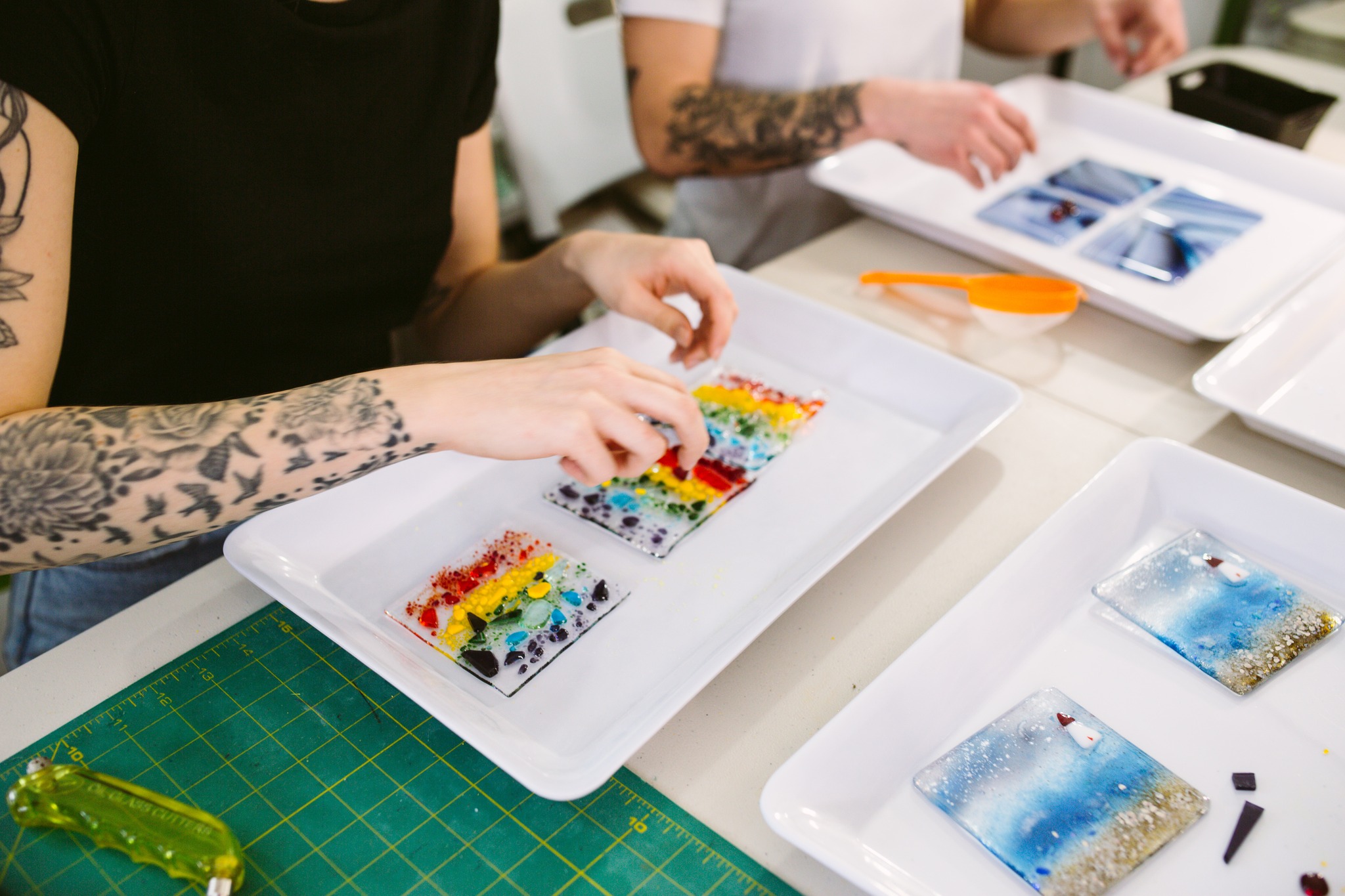 two people arranging vibrantly coloured glass blocks into their glass projects, which rest on white rectangular plates on a tabletop. 