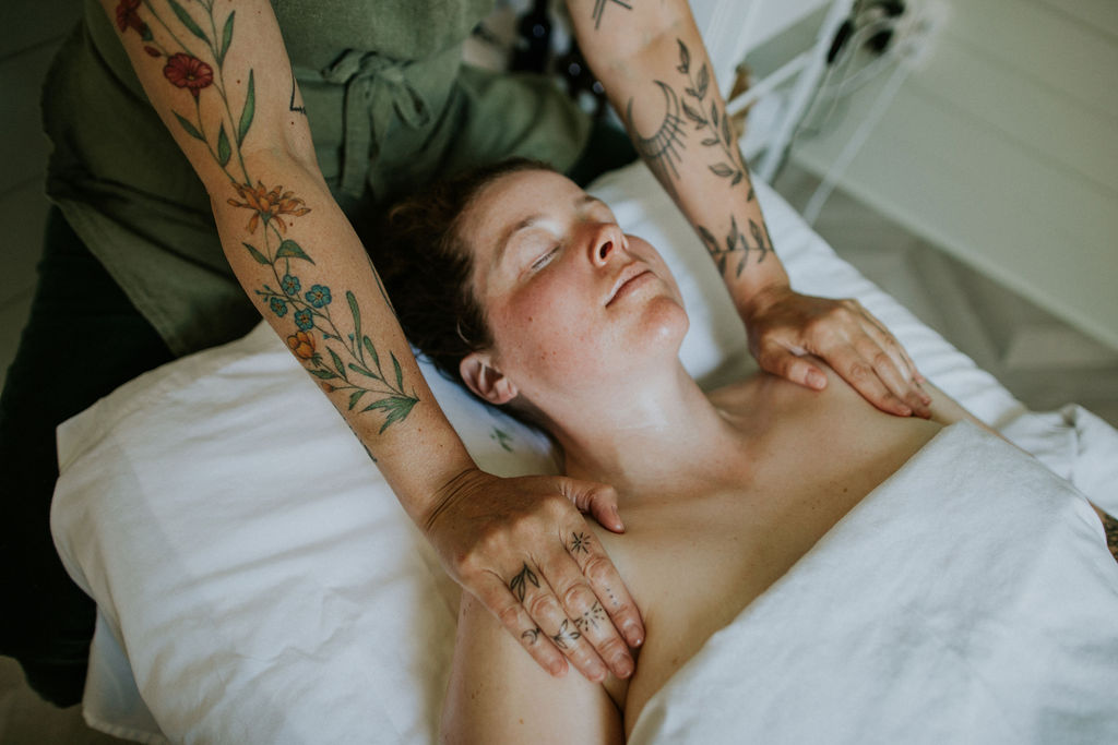 A woman with beautiful floral tattoos on her arms massages the shoulders of a smiling woman in a white spa bed