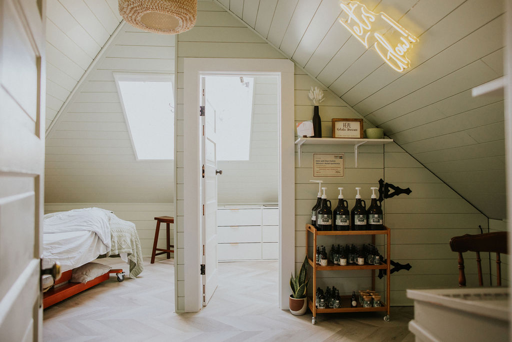 A white and brightly-lit, high-roofed healing room, with a sign the says "Let's Glow". There is a shelf of green bottles filled with beauty and healing products.