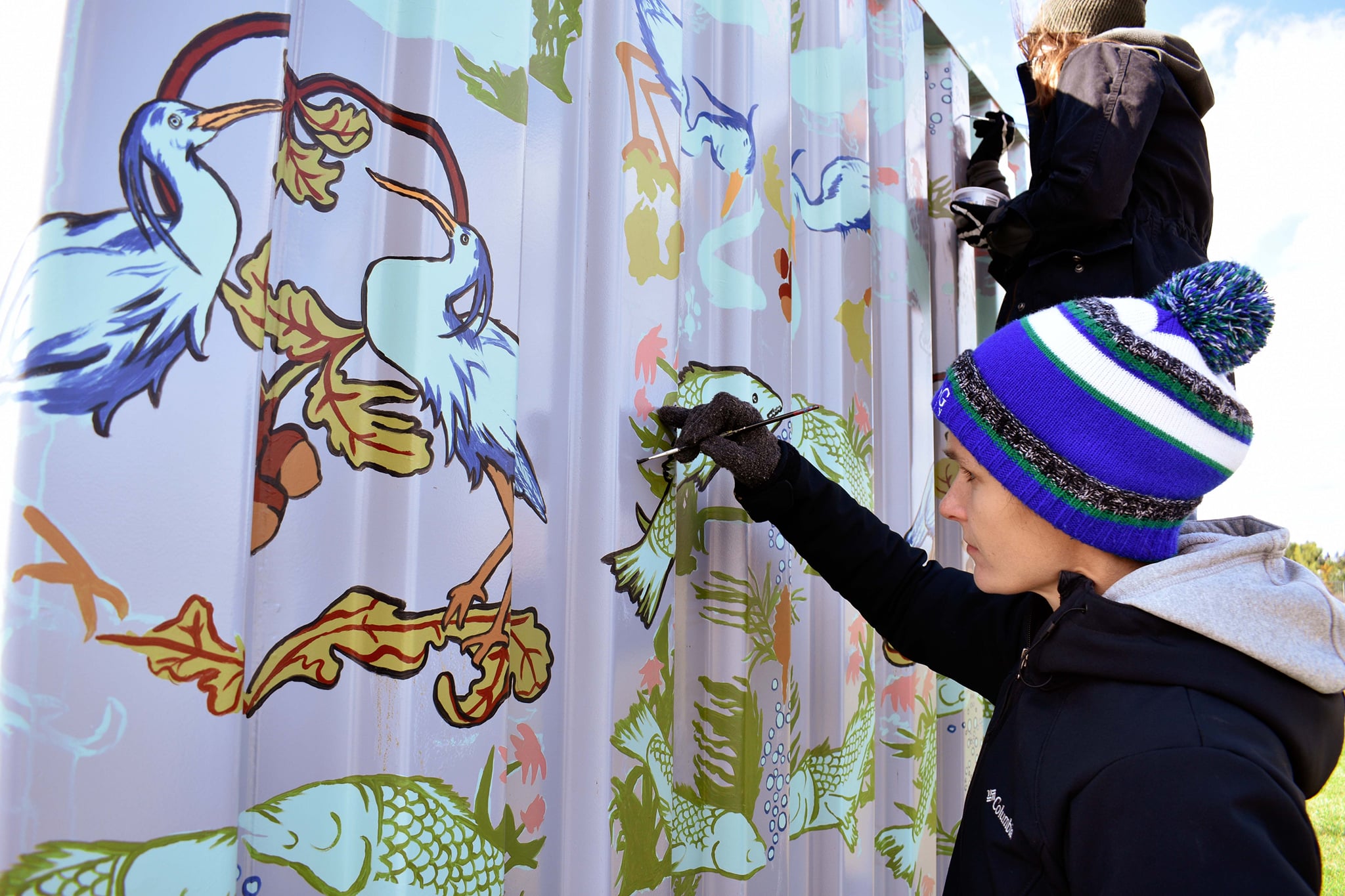 artist Laura Peturson, wearing a black coat, gloves and blue striped toque, painting a beautiful bird and floral motif on an outdoor wall.