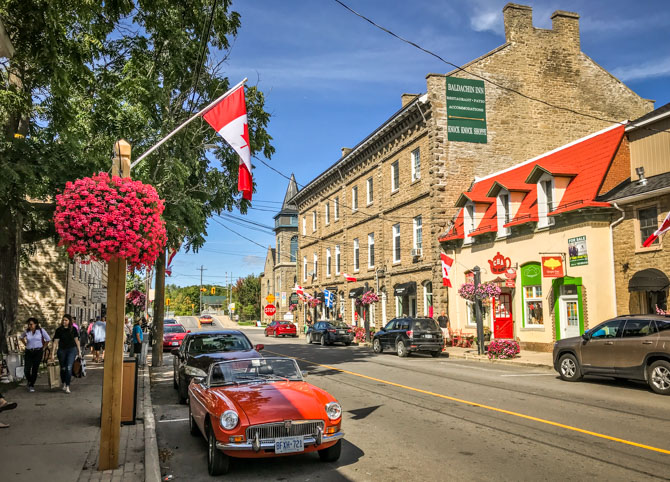 a red antique car parked on a clean Merrickville street, lined with potted flowers, trees, and attractive old stone buildings under a bright blue sky.