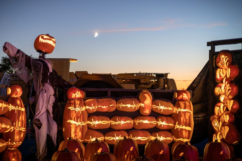 Glowing jack-o-lanterns stacked to resemble a barbed-wire fence, next to a scarecrow with a lit jack-o-lantern head, under a darkening sky with crescent moon