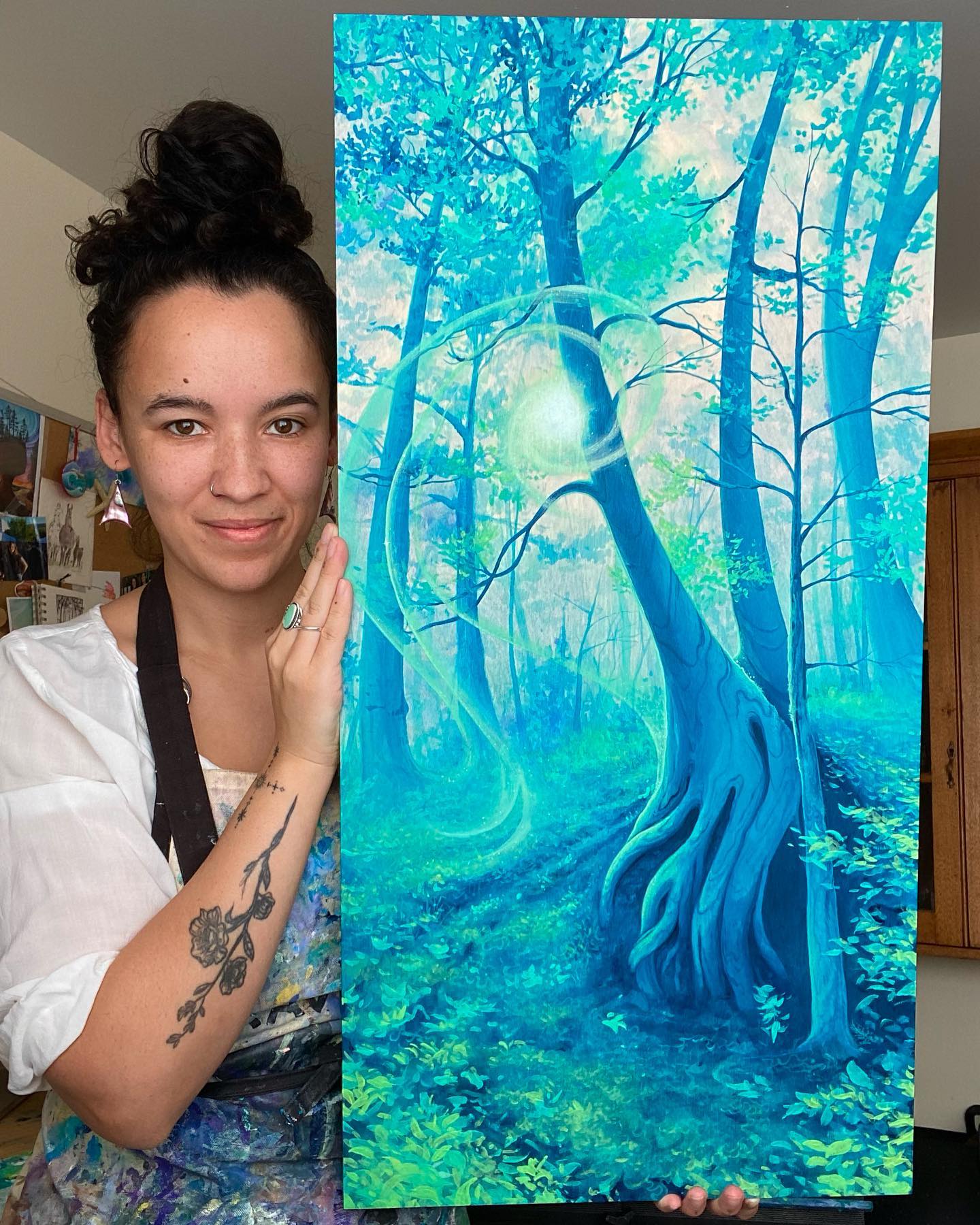 Penny Heather holding a painting titled "Reminiscence". It shows a forest with sun behind the trees, painted in blue and aqua tones in a dreamlike effect.