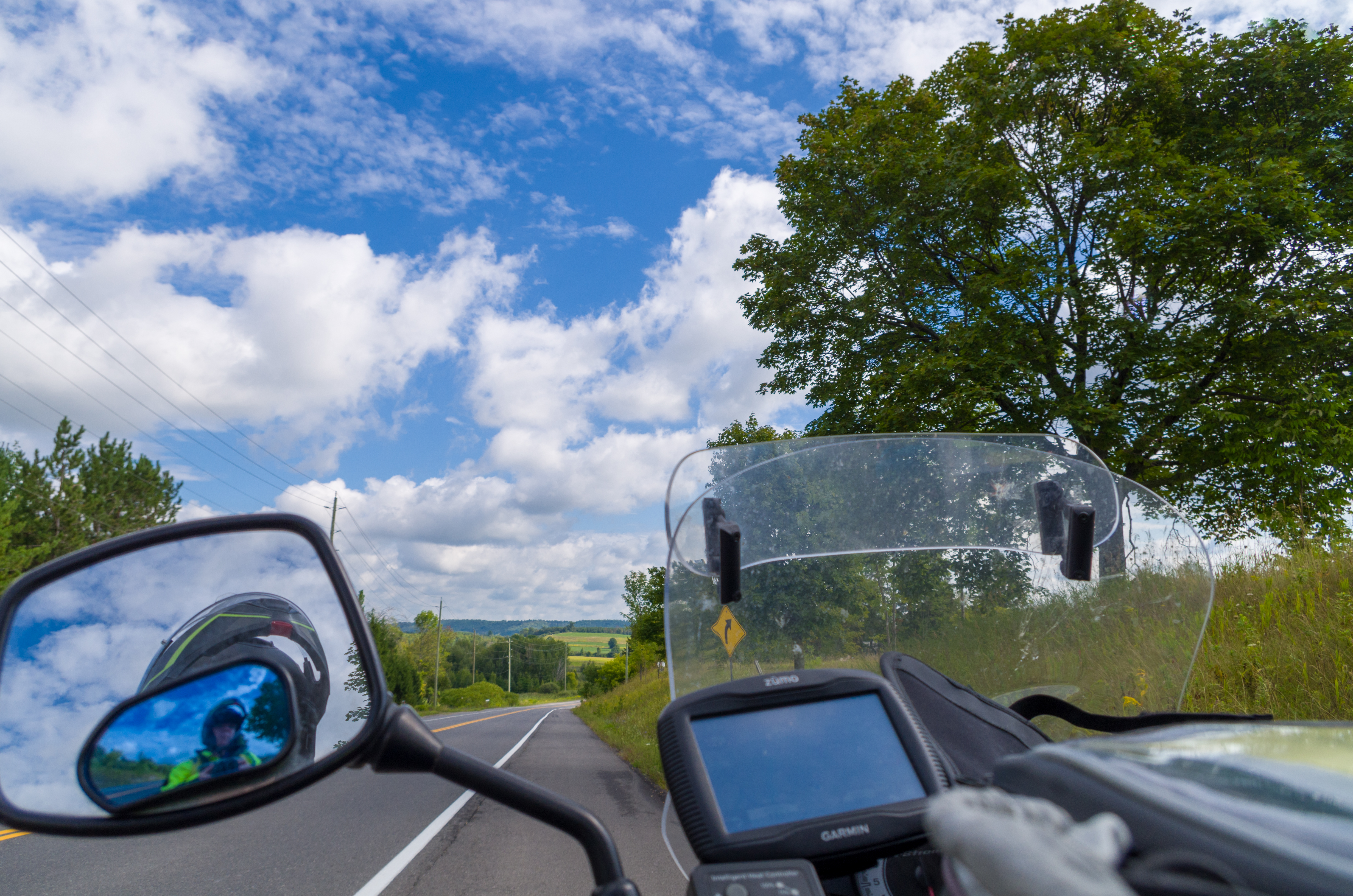 Blue sky, green trees and fields and an open road, seen over a motorcycle rearview mirror
