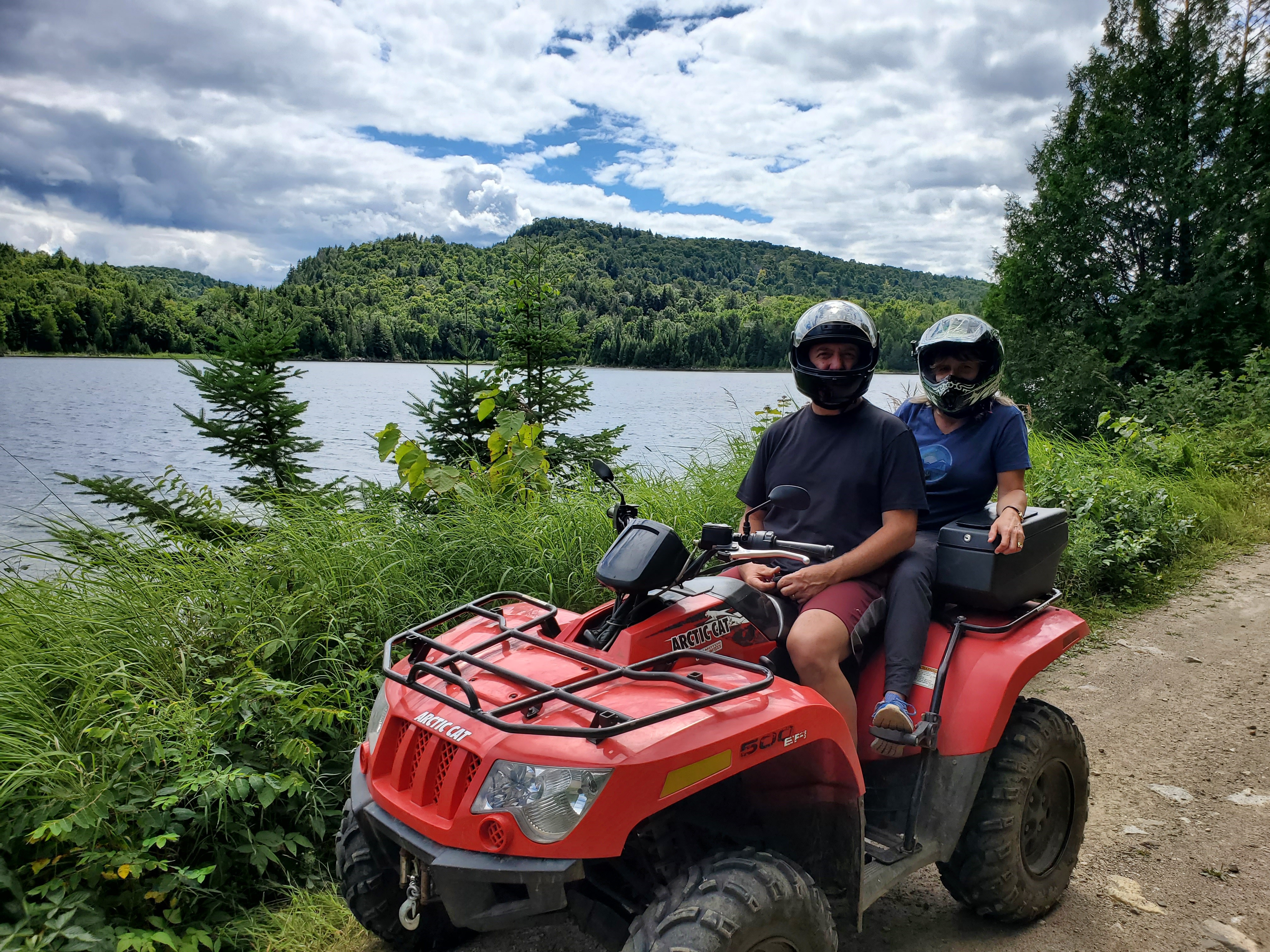 two people wearing helmets and sittting on a red ATV, in front of lush green forest and a lake.