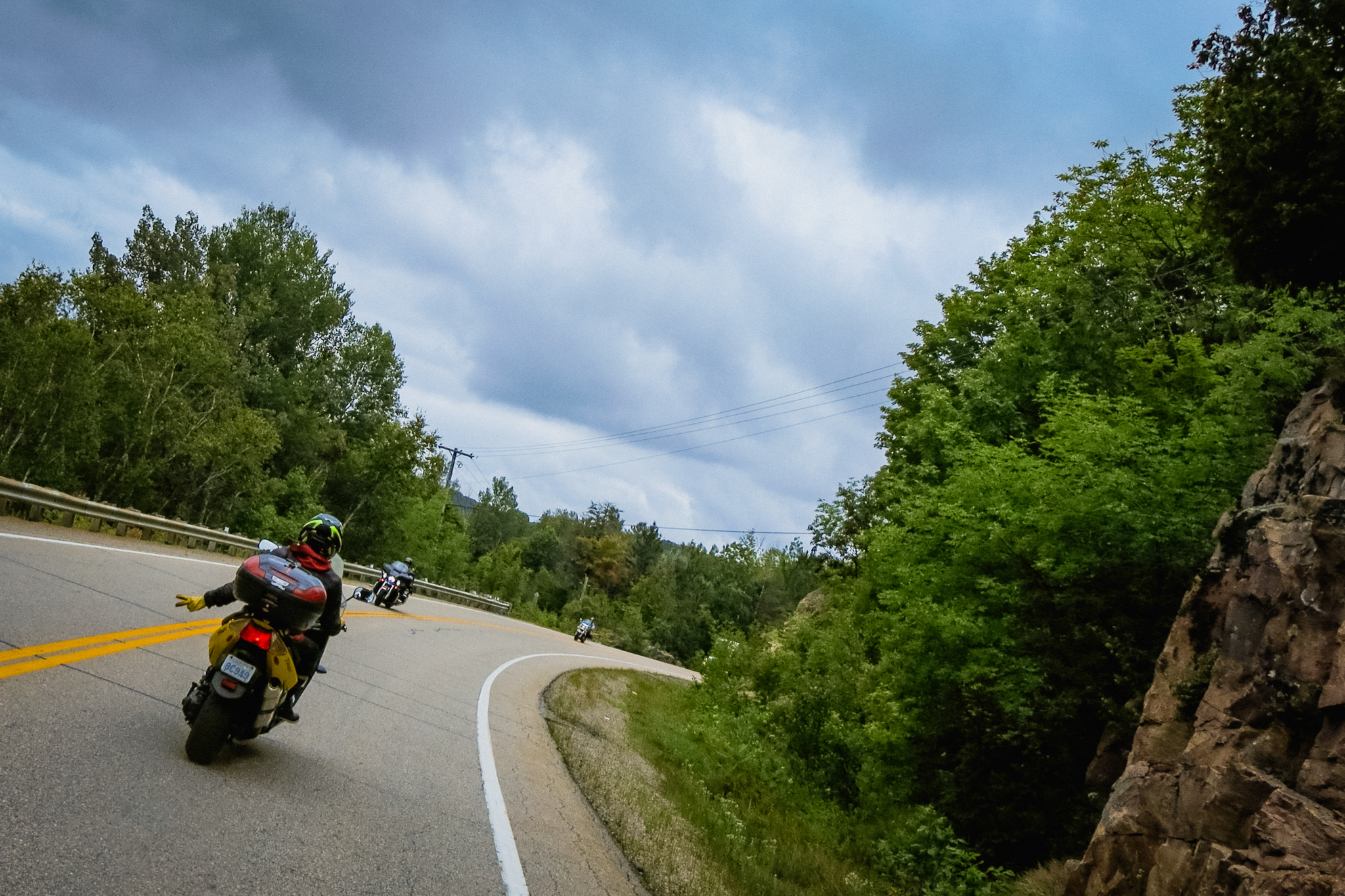 A motorcyclist from behind, giving a two-fingered wave to a passing biker. They are rounding a corner on a quiet highway surrounded by lush green forest and under blue-grey storm clouds.