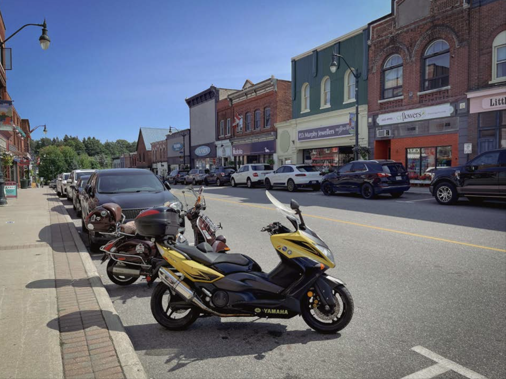 two motorcycles parked along Manitoba Street in Bracebridge; a tidy-looking street lined with attractive antique buildings, under a bright blue sunny sky.