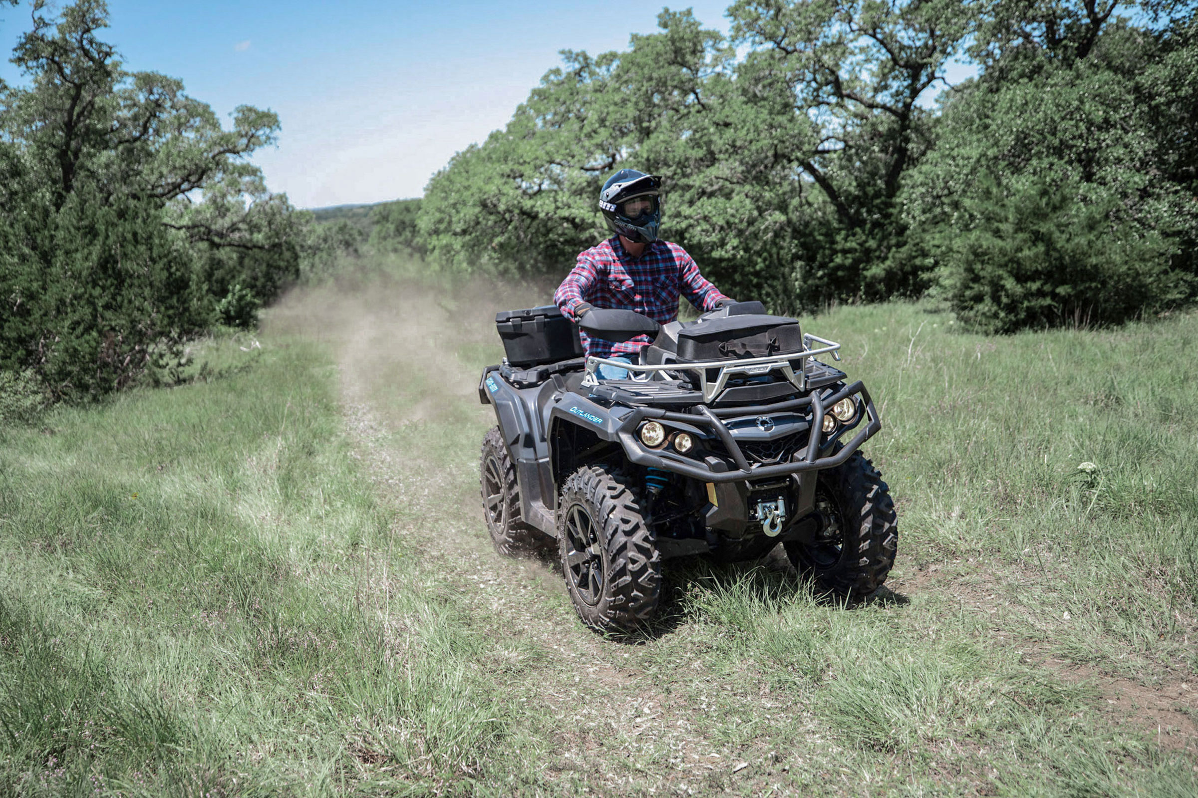 A rider on a new Can Am Outlander ATV, riding on a grassy trail lined with lush green trees under a bright blue sky