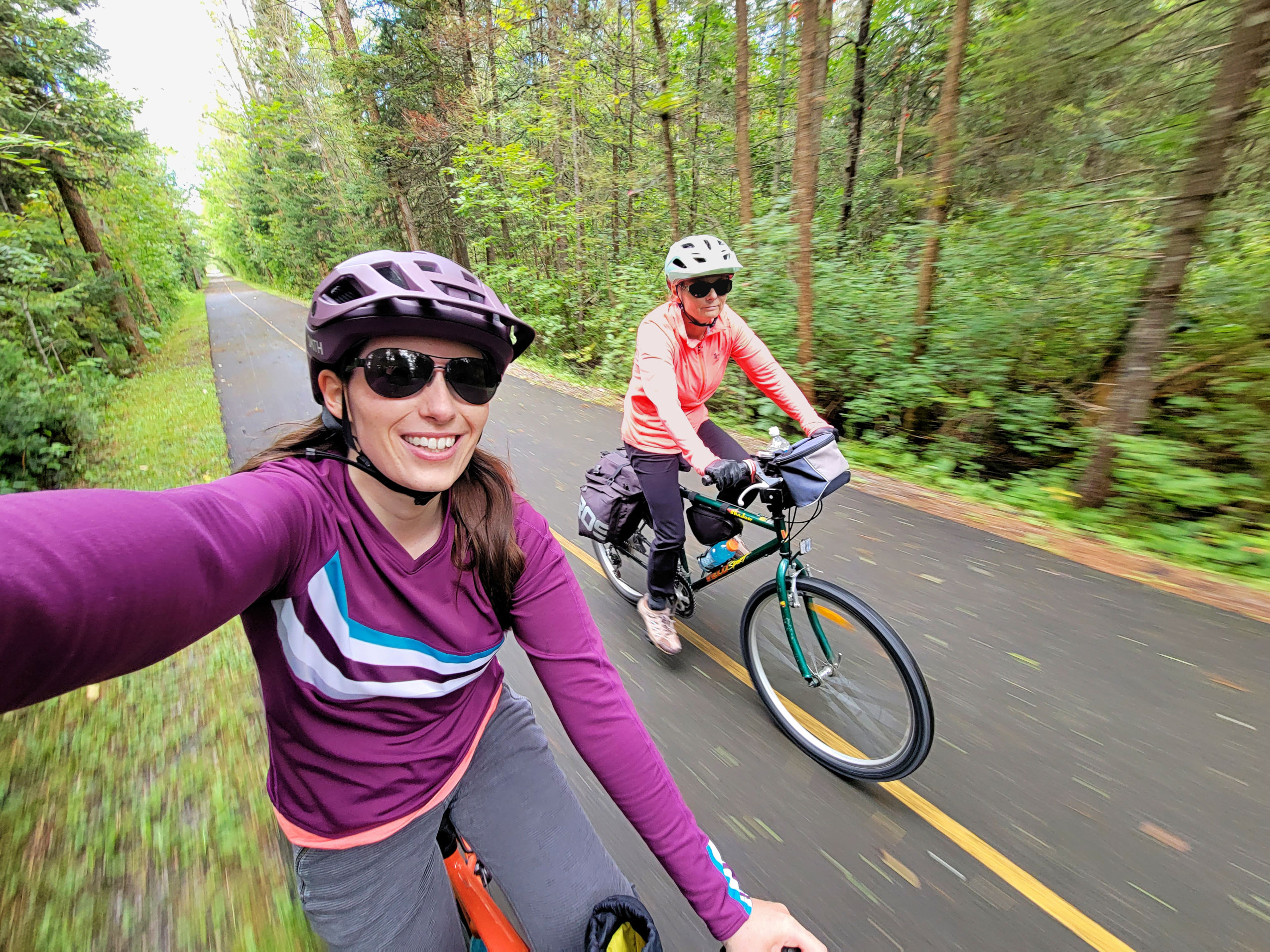 woman takes a selfie while riding a bicycle along a paved forest trail