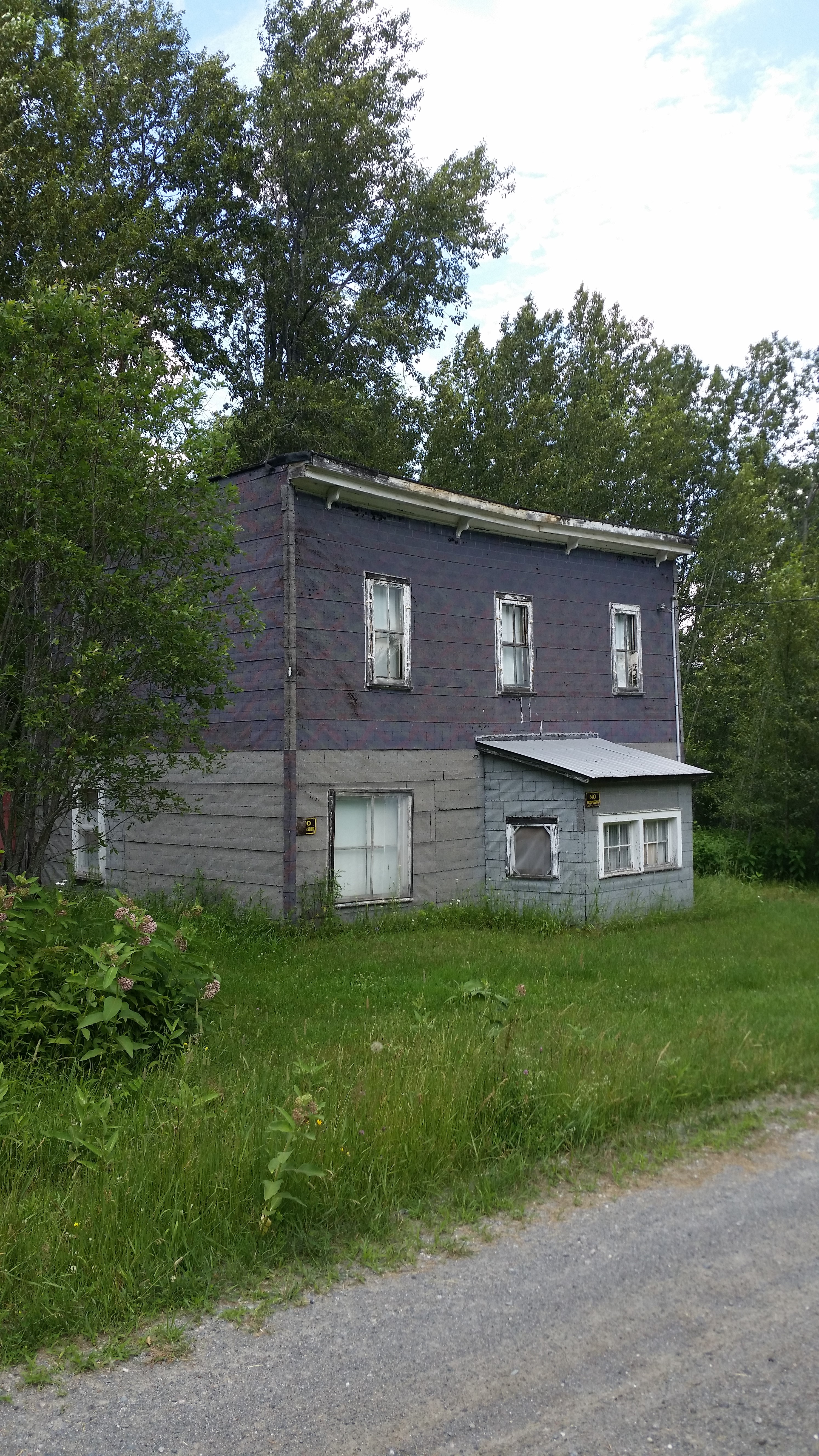 the old Desaulniers Hotel; an abandoned grey, early-1900s building with two levels and many simple windows, surrounded by grass, trees and a gravel road.