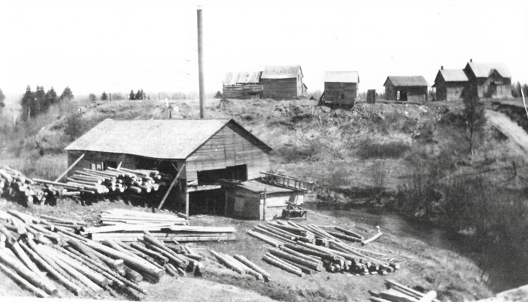 an old black and white photo of the Desaulniers sawmill; several small, simple wooden buildings surrounded by small trees and brush, and a lumberyard full of stacks of long logs.