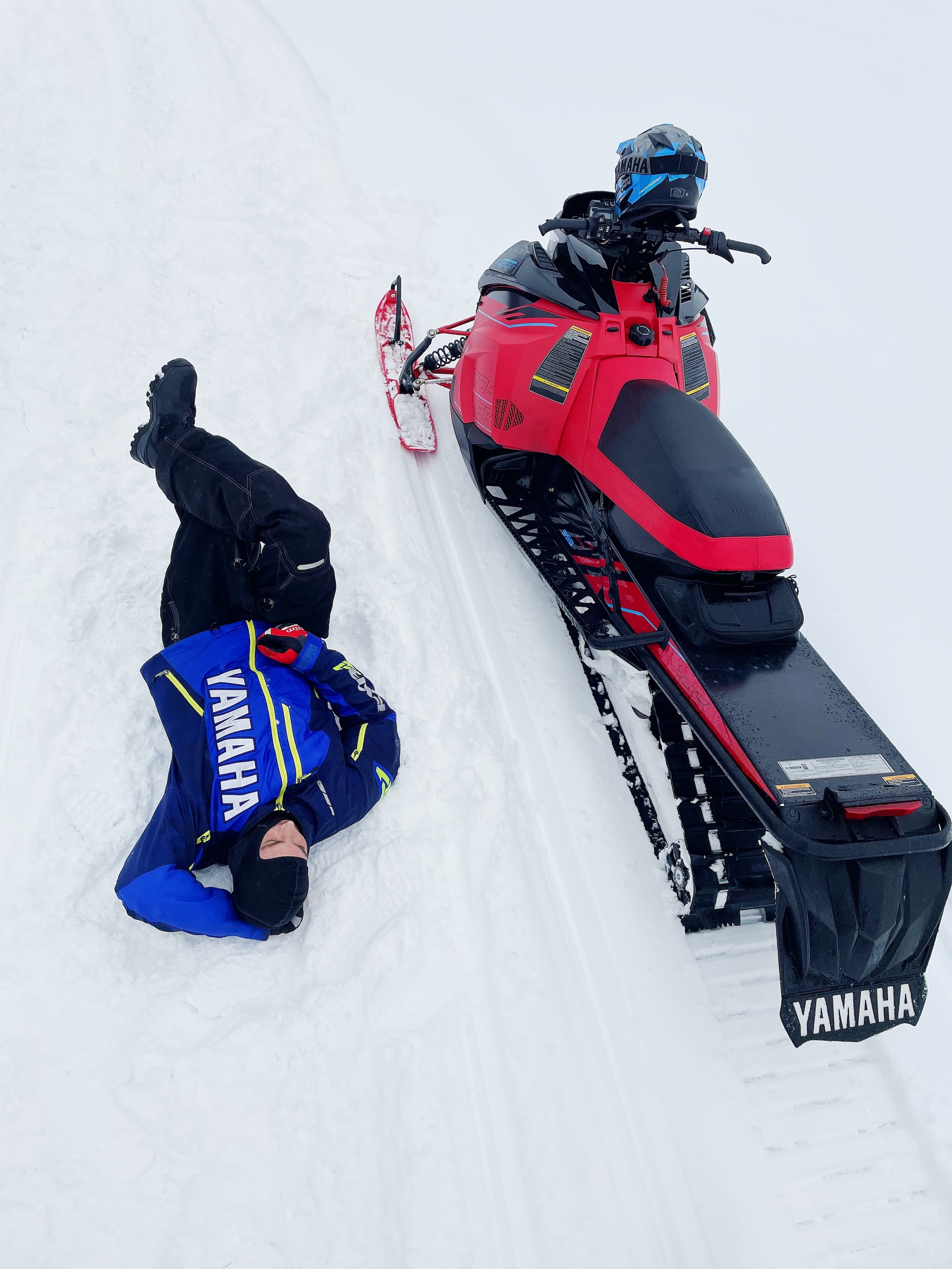 a shot from a above showing a man laying in a relaxed pose in the snow, next to his snowmobile.