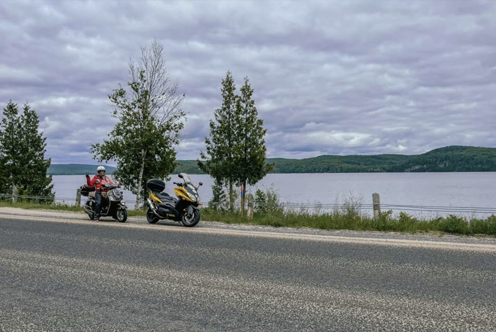 two motorcycles, one with waving rider, parked along the side of the road next to a large, calm lake surrounded by green forest, under a stormy grey sky.