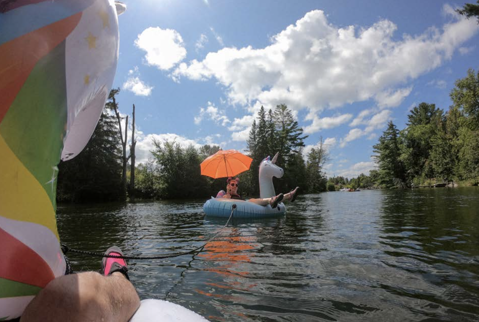 a person wearing sunglasses floating down a river in a large inflatable unicorn floaty, shaded by an orange umbrella. The leg of the photographer can be seen, as well as the head of another inflatable unicorn that they sit in.