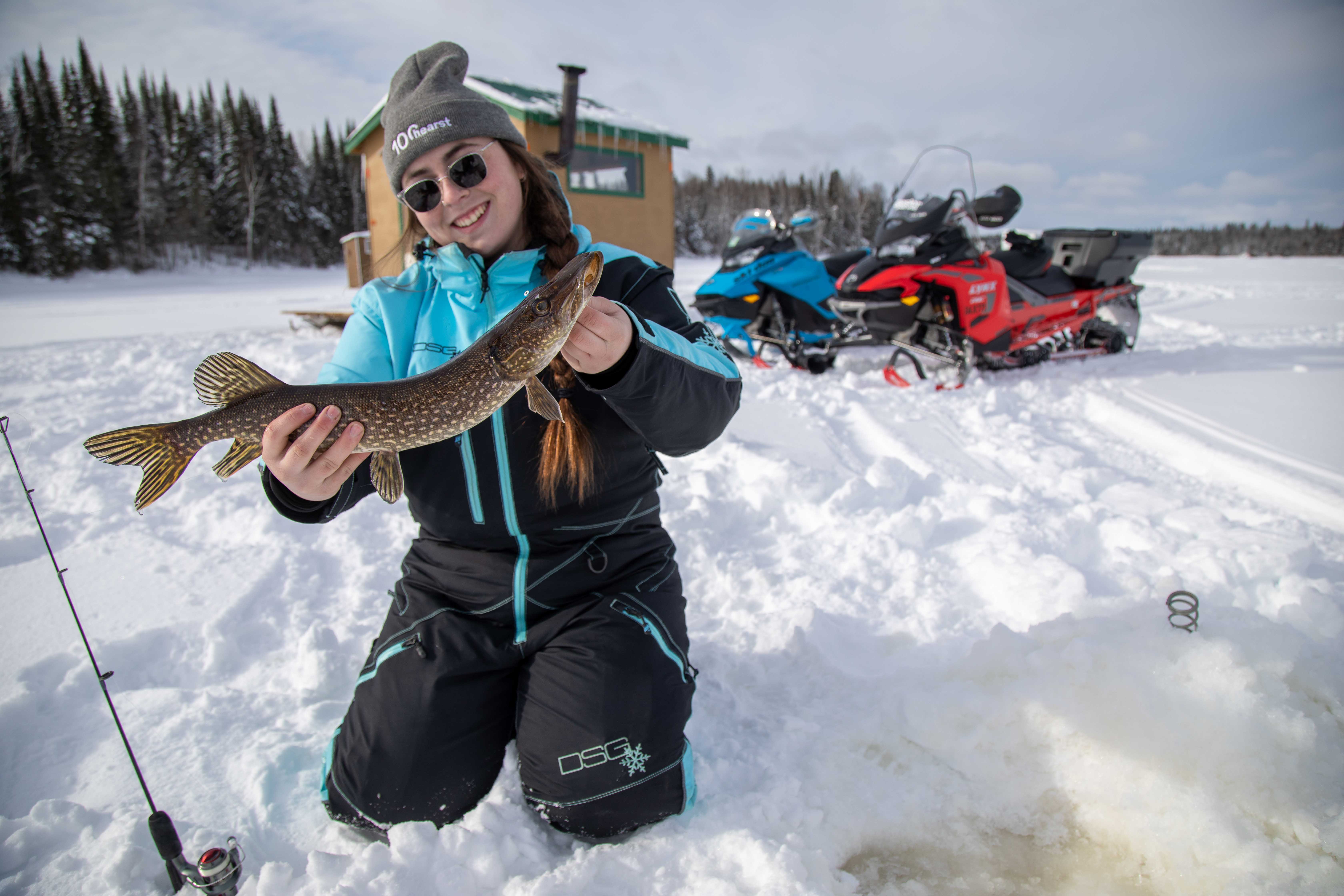 a smiling woman in snow gear kneeling next. to an ice fishing hole, holding a large fish. 2 snowmobiles are parked in the background.