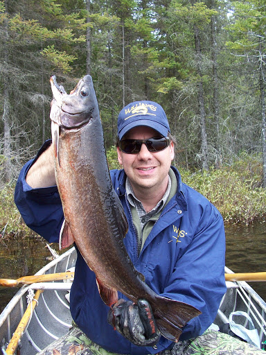 a smiling man in blue coat and sunglasses holding up a large aurora trout. Green spruce forest and lake water are in the background.