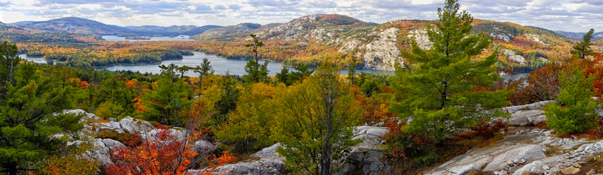 a view from Killarney Provincial Park; smooth rock mountains and a valley dappled with green, red and yellow forest with a lake in the distance. 