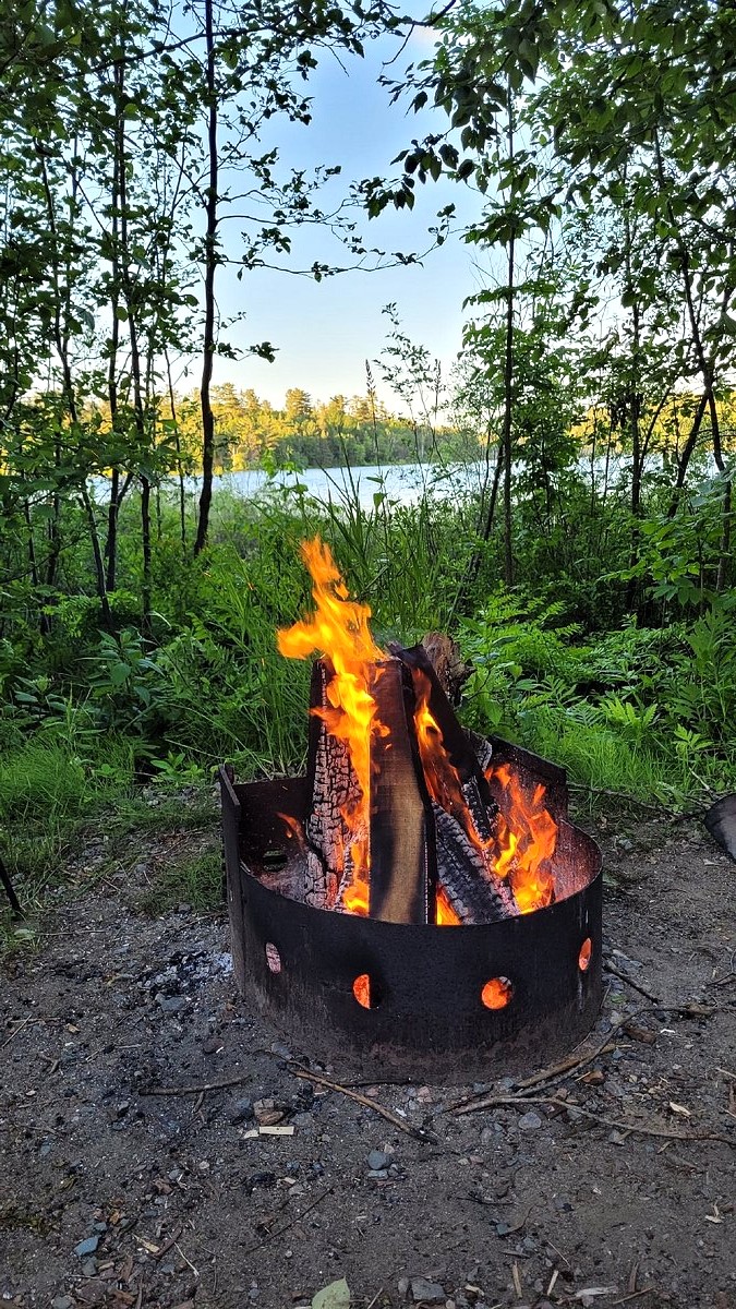 a metal firepit with roaring orange fire, in front of bright green grass and forest. Through the trees there is a blue lake, bright blue sky and late afternoon sun.