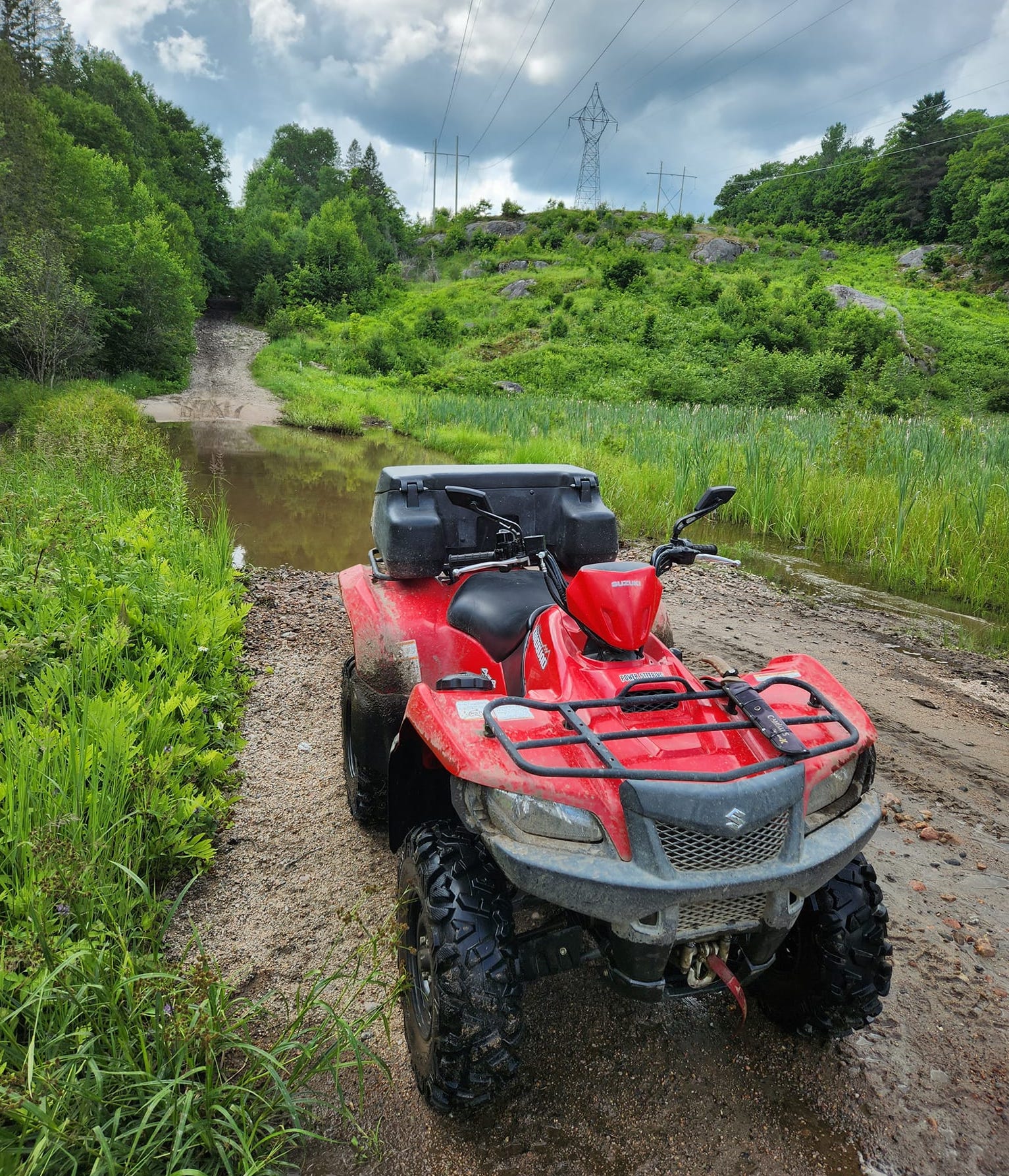 a red ATV parked on a dirt trail, surrounded by lush green forest. A large section of the trail in the background is flooded with water. 