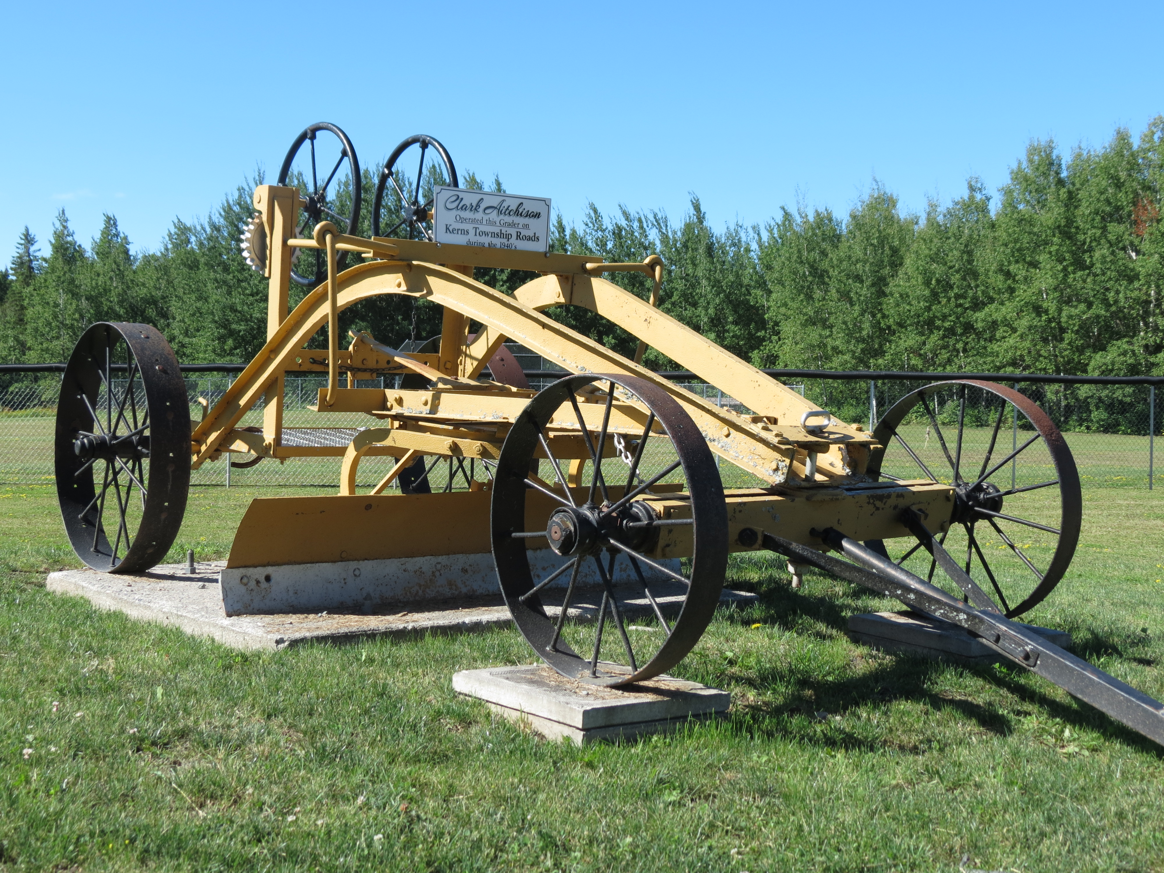An antique yellow metal road plough on 4 iron wheels, designed to be pulled behind horses. It is displayed on the Milberta Church grounds, surrounded by green grass and lush green trees, under a bright blue sky.