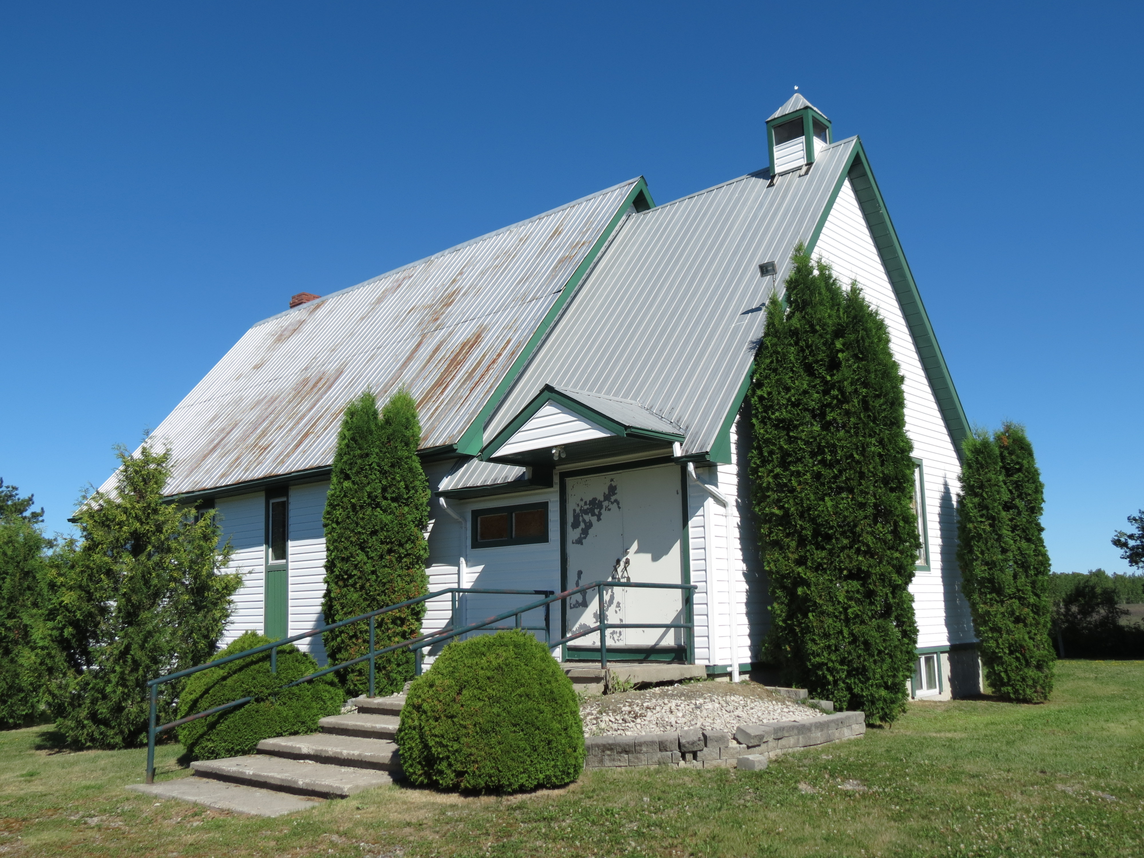 The Milberta Church; a large white and green wooden church from the early 1900s, slightly weathered and surrounded by tall green manicured shrubs and green grass 