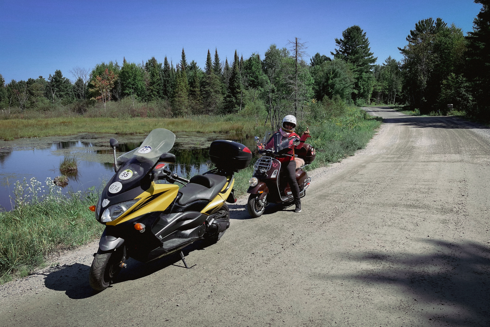 two motorcycles, one with a rider, parked along the side of a road next to a pond surrounded in green grass and forest, under a bright blue sky.