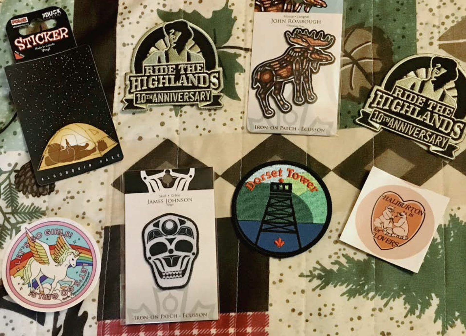 a collection of embroidered patches laid out on a bedspread printed with evergreen trees. Some of the patches advertise "Ride the Highlands", "Dorset Tower", and a rainbow pegasus patch the says "Let's go Girls".