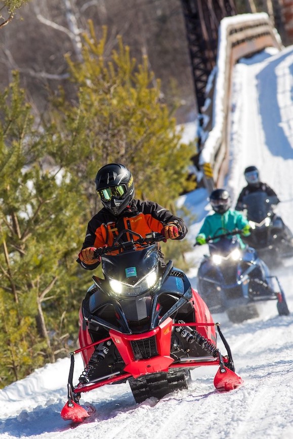 a line of three people on snowmobiles riding down a snowy trail, a snow covered wooden bridge and green spruce trees behind them.