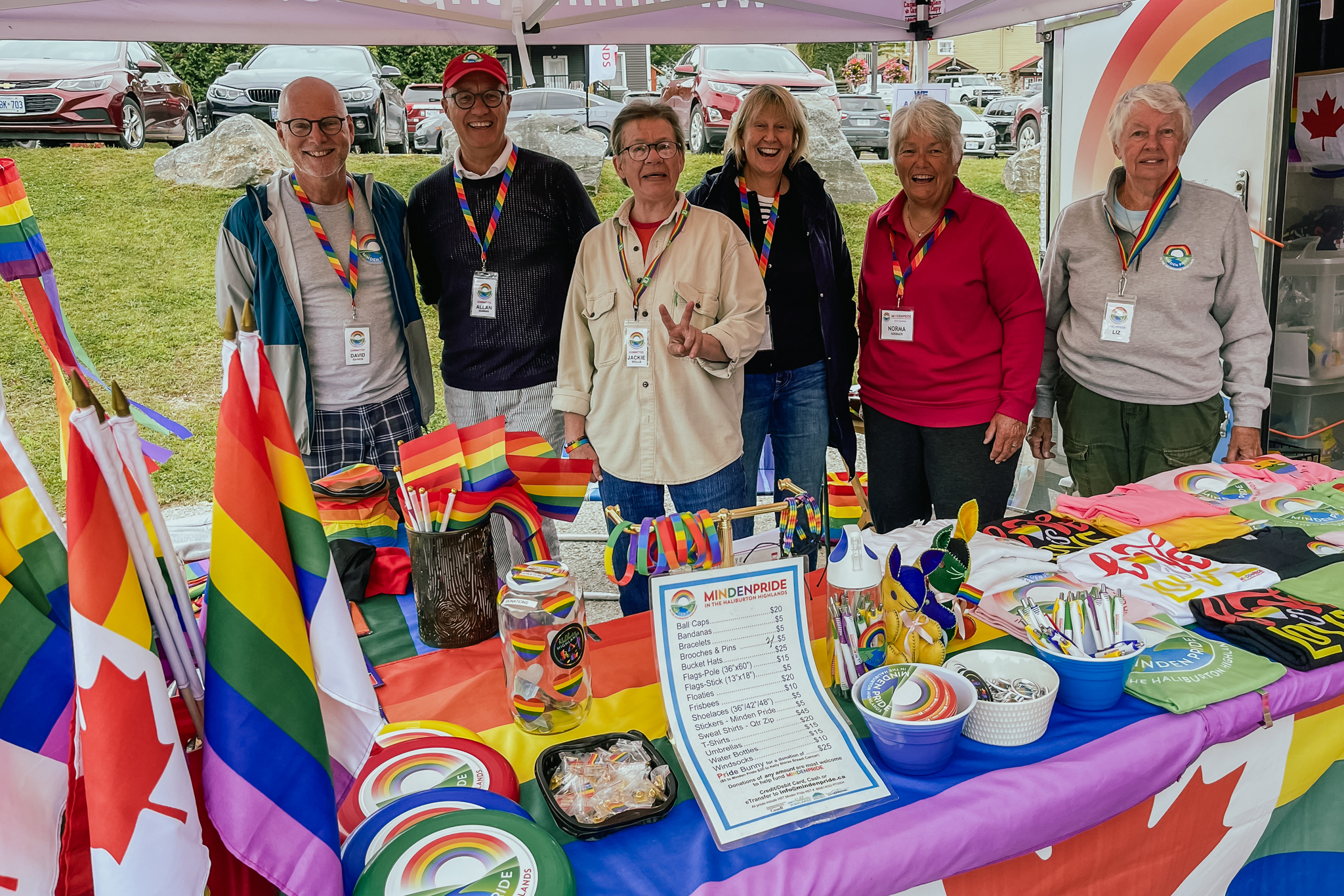 6 smiling volunteers with badges hanging around their necks pose behind a display table full of rainbow flags and inspirational memorabilia