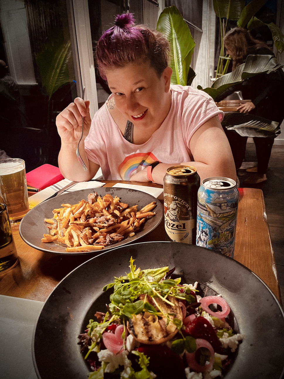 a smiling person with a pink shirt and purple hair sitting at a restaurant table, holding a fork over a giant plate of poutine and a salad
