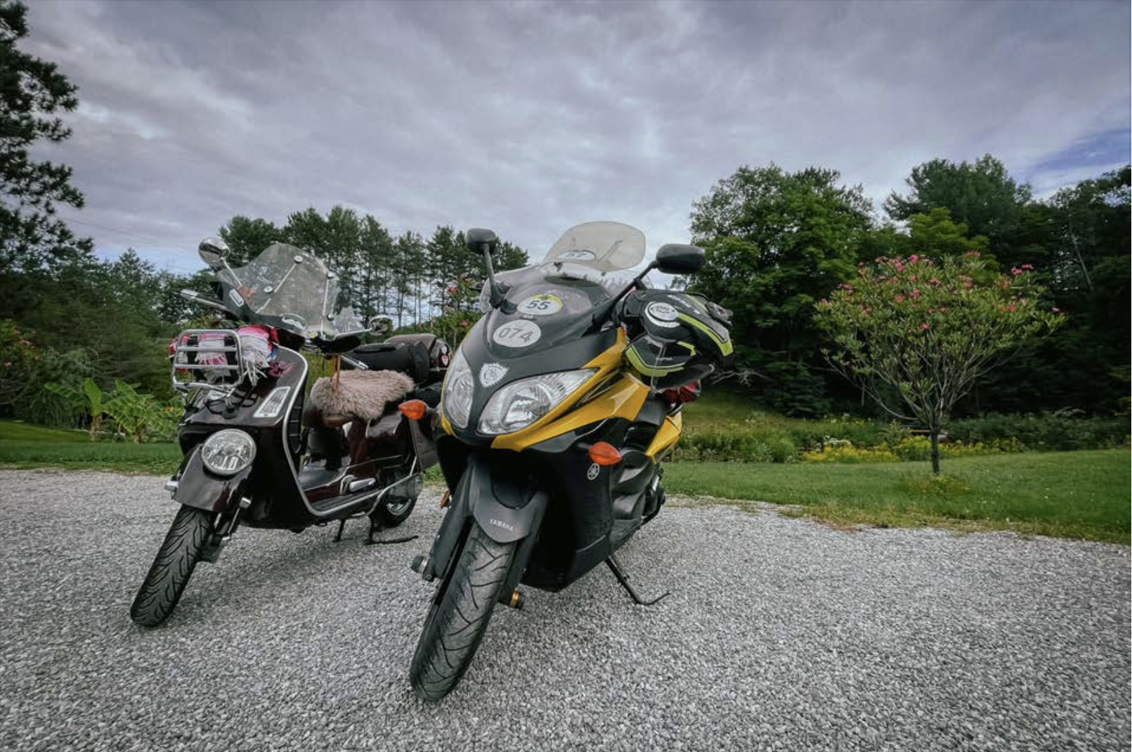 two motorcycles parked side by side on gravel, with lush green grass and trees and stormy grey clouds in the background.