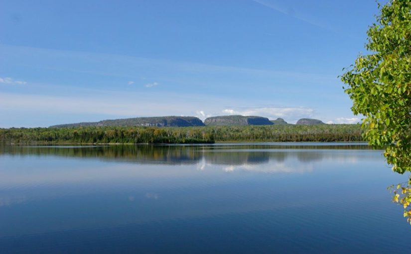 Sleeping Giant; a large, glassy blue lake with a forested peninsula on the other side, under a bright blue sky