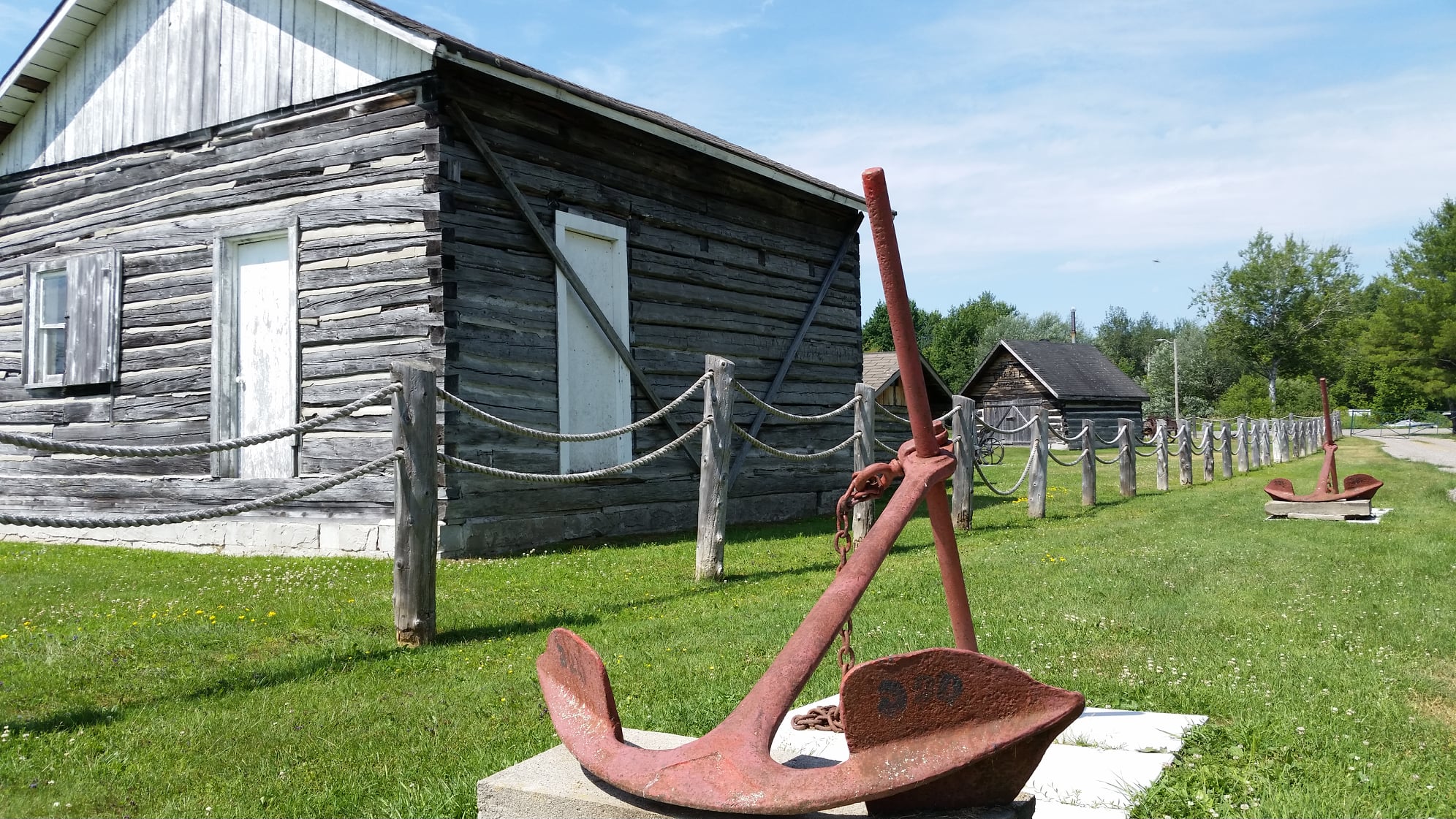 Sturgeon River House Museum; a log building surrounded by a wood and chain fence and green grass and trees, with a display of old, rusted metal tools in the foreground.