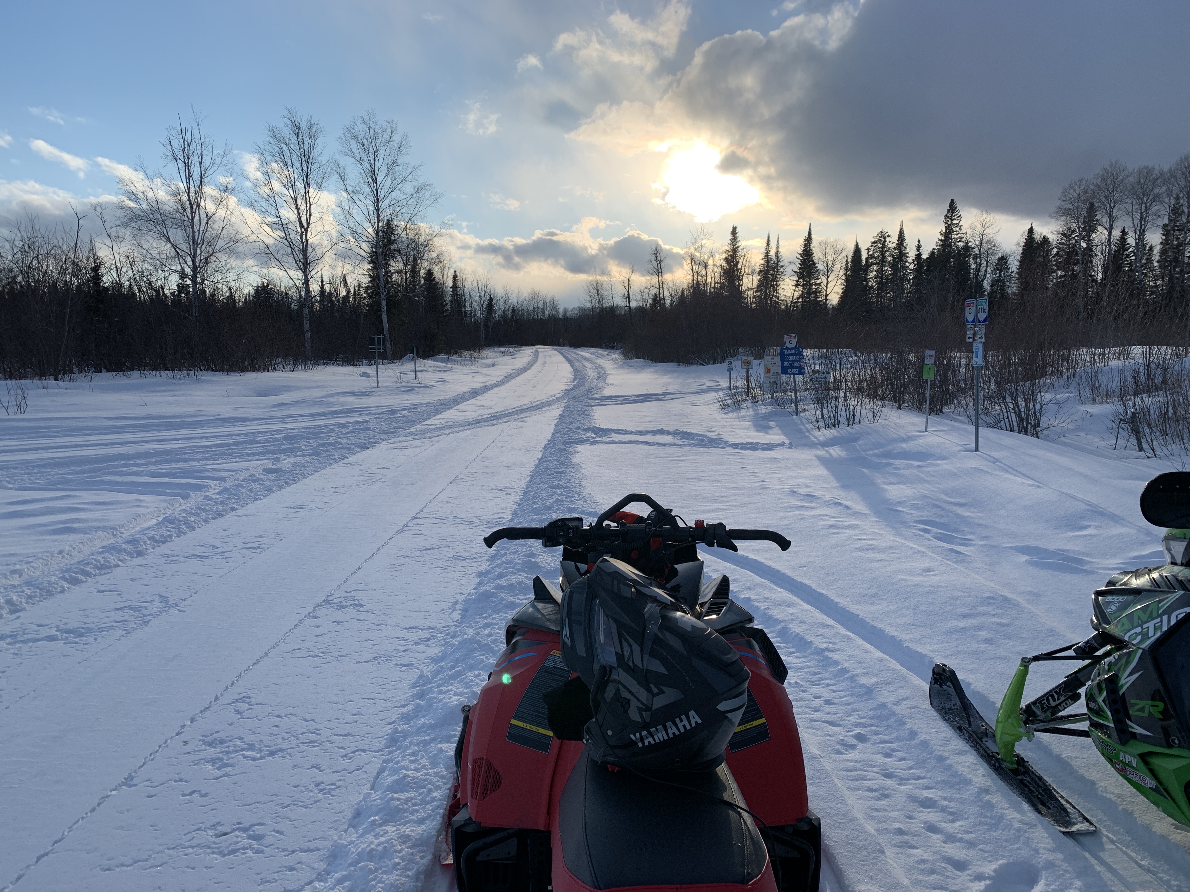 two snowmobiles facing a sunrise with storm clouds rolling in, on a snowy forest trail
