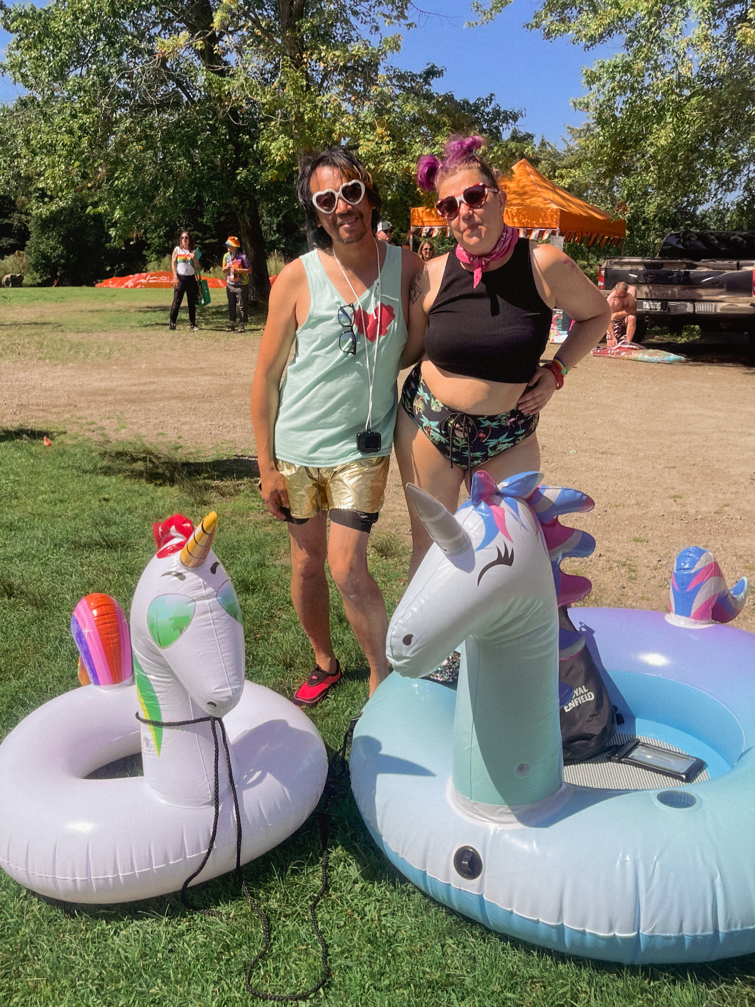 two smiling people with arms around each other's shoulders, wearing sunglasses and bathing suits and standing next to two large inflatable unicorn floaties. They are surrounded by green trees and grass under a bright blue sky. 