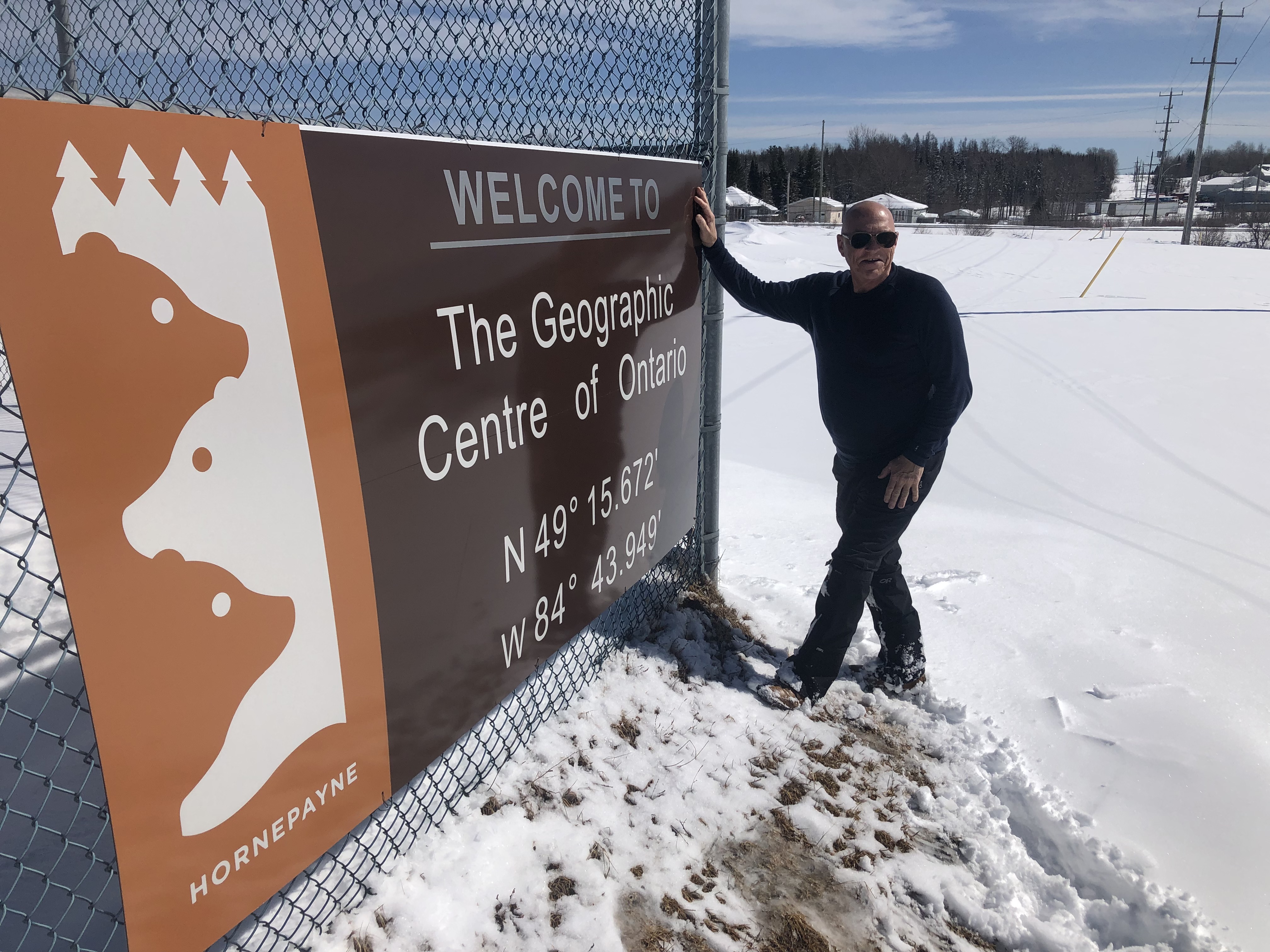 Bill Steer wearing a black jacket and pants, standing next to a large sign tnat says "Welcome to the Geographic Centre of Ontario". The ground is snowy and the sky is blue.