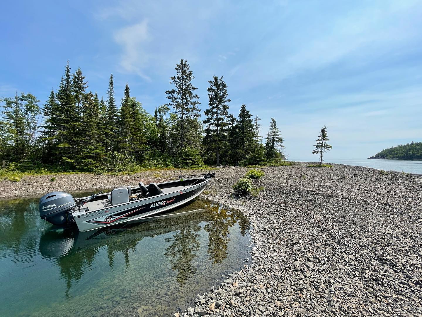 a silver motorboat beached in glassy green lake water at a pebble shorline, under a bright blue sky. Green conifer trees are in the background.