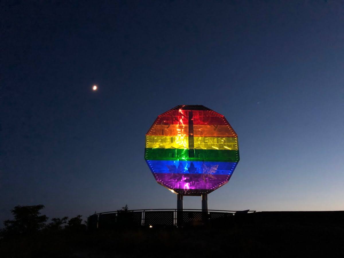 Sudbury's Big Nickel monument in front of a night sky with a single star to the left; rainbow stripes are projected onto the giant coin's front, lighting it up and making it appear to be painted with rainbow stripes.