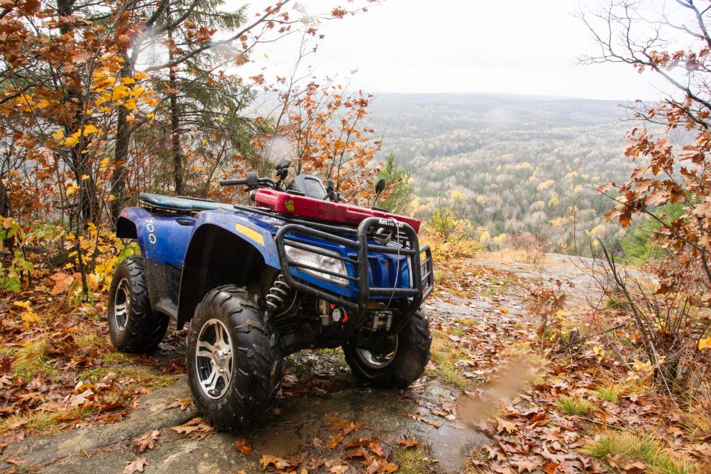 A blue ATV parked on a forested cliff, overlooking a wide forested valley. All the trees are shades of orange, yellow and gold.