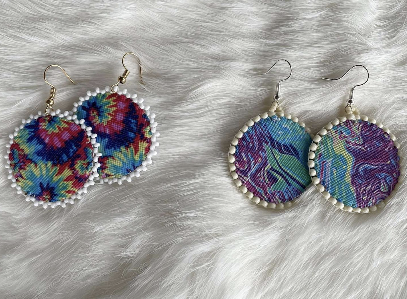 two sets of round, handbeaded earrings in purple, turquoise and green tones on a white fur background.